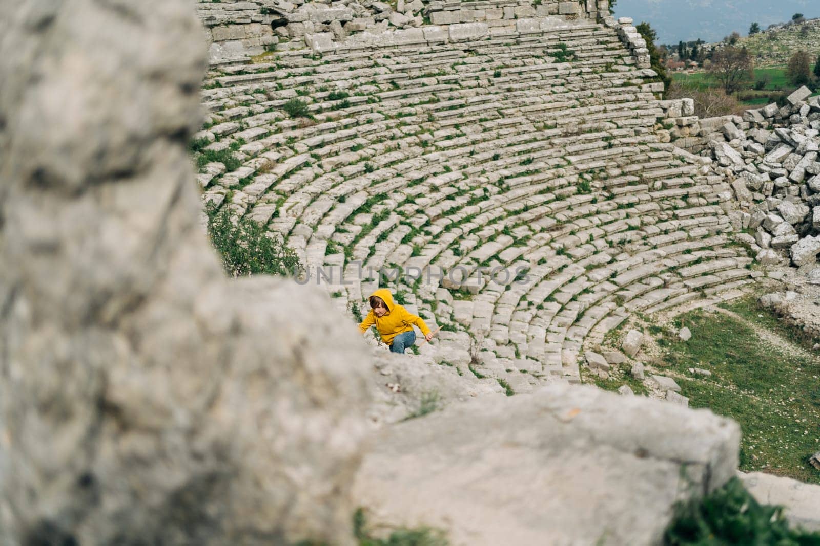 School boy kid in yellow hoodie and blue jeans exploring remains of ancient arena stone coliseum amphitheater by Ostanina