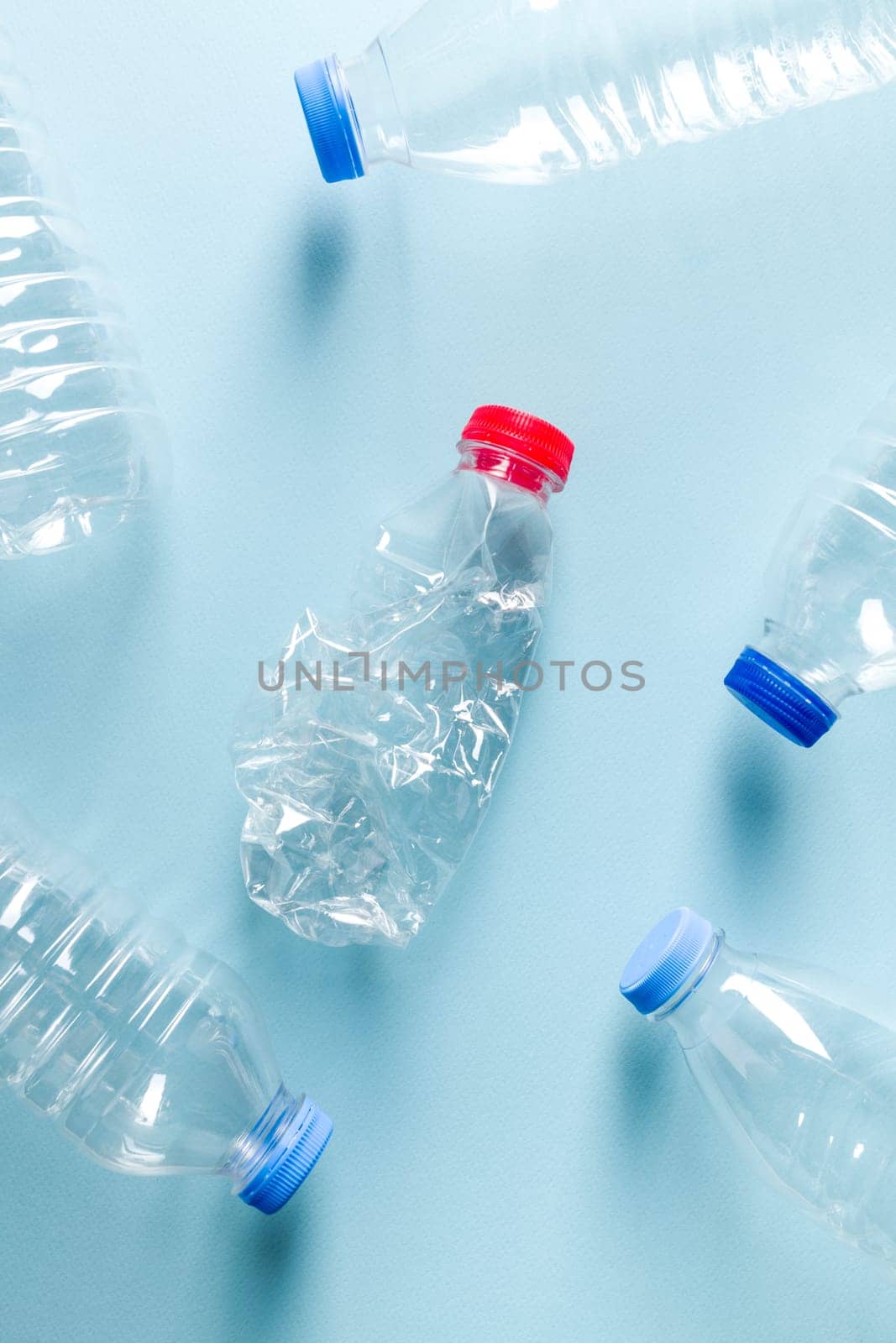 Crushed plastic water bottles with blue caps standing around crushed plastic water bottle with red cap on blue background by Sonat
