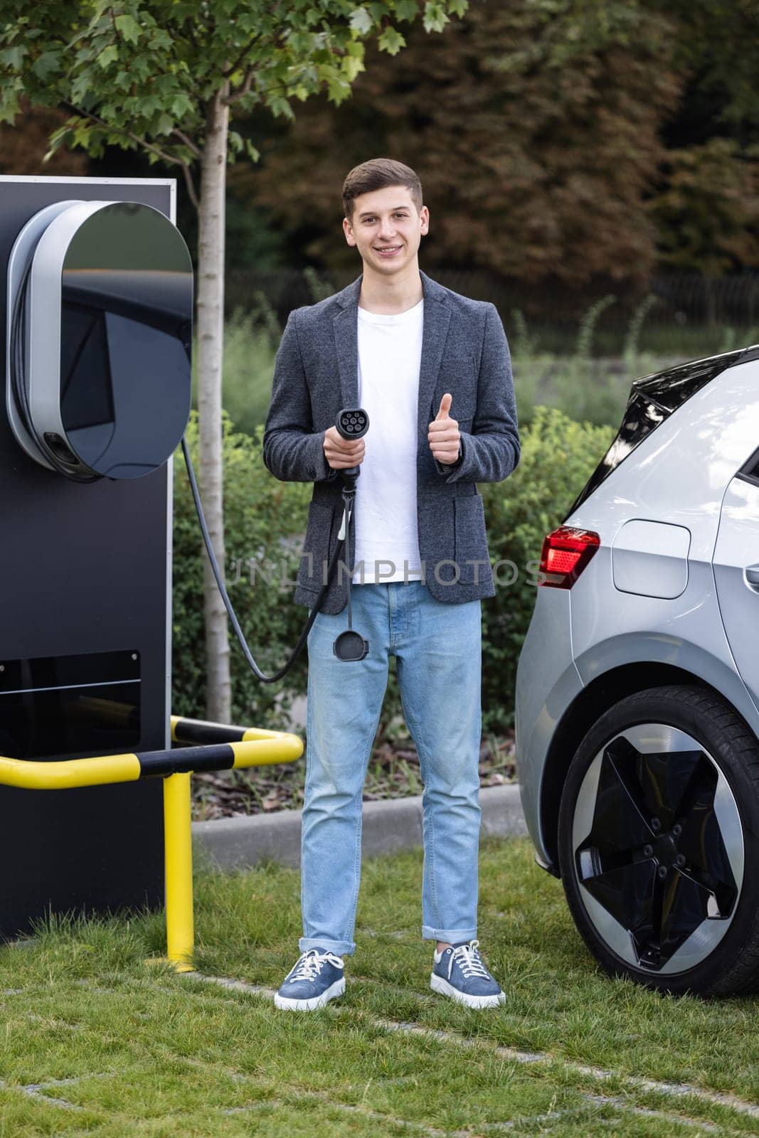 Caucasian man standing near electric charging station looking at camera and showing thumb up. Male holds a charging cable in his hands. Eco-friendly modern green transport by uflypro