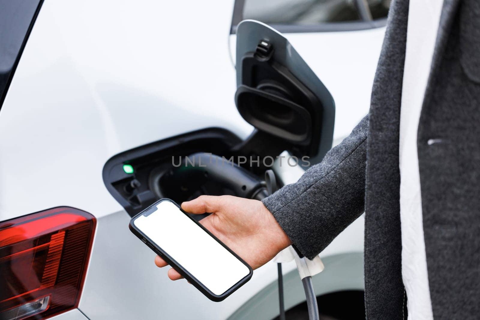 Smartphone monitoring of charging. Young man with her electric car at daytime. Man holding smartphone while charging car at electric vehicle charging station, closeup.