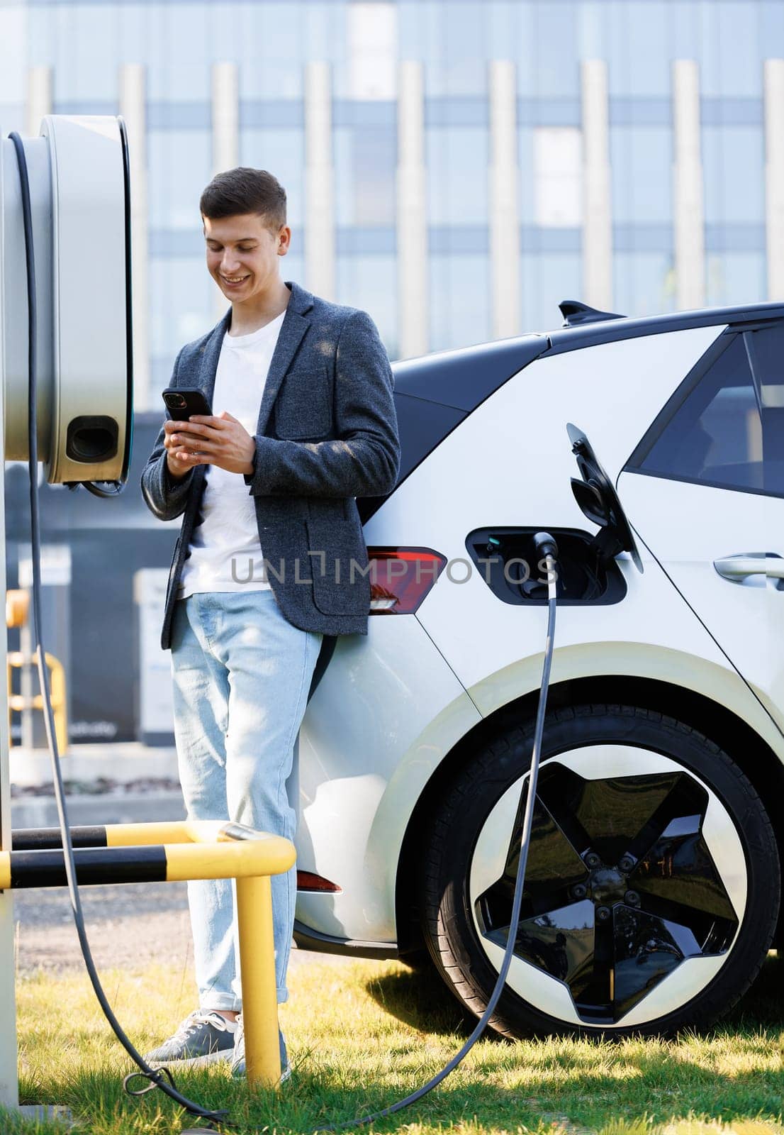 Young man using smartphone near electric car on charging station. Man holding smartphone while charging car at electric vehicle charging station. Businessman standing in background and waiting.
