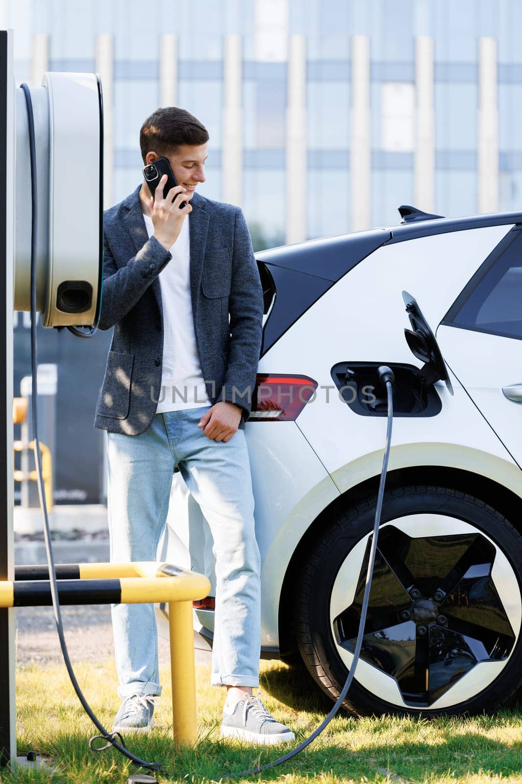 Handsome businessman calling while charging his electric car on the street. Portrait of joyful 25-aged man which has mobile conversation while electric car charging battery on charging station by uflypro