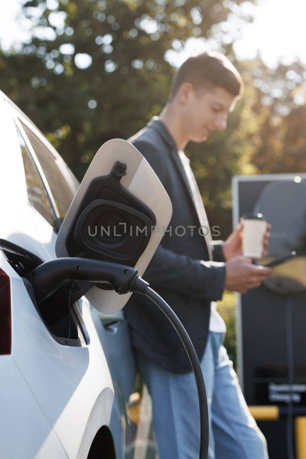 Caucasian man using smartphone and waiting power supply connect to electric vehicles for charging the battery in car. Plug charging an electric car from charging station by uflypro