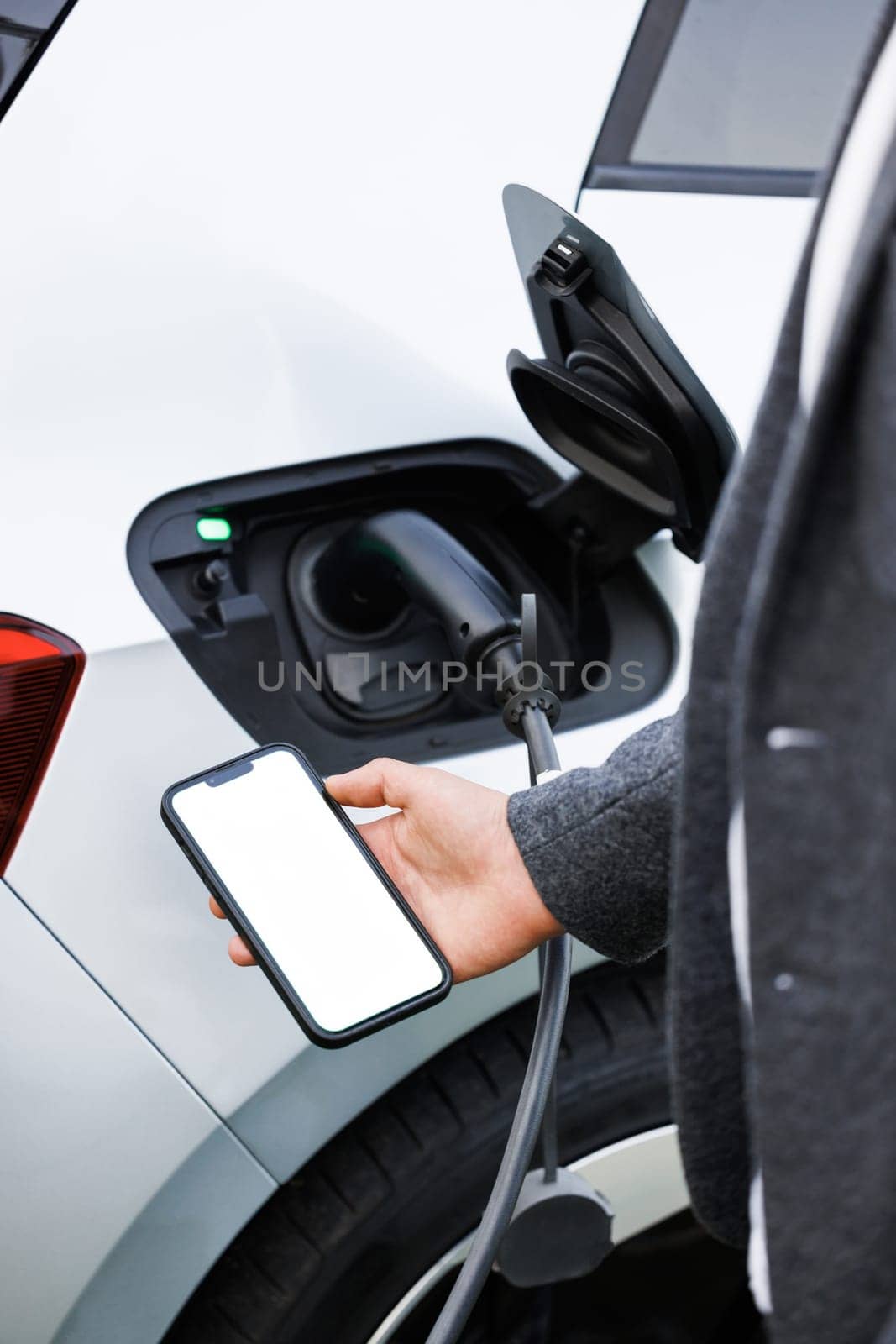 Businessman traveling by electric car having stop at chraging station standing holding smartphone looking at screen copy space for text or product close-up while having vehicle fully charged. by uflypro