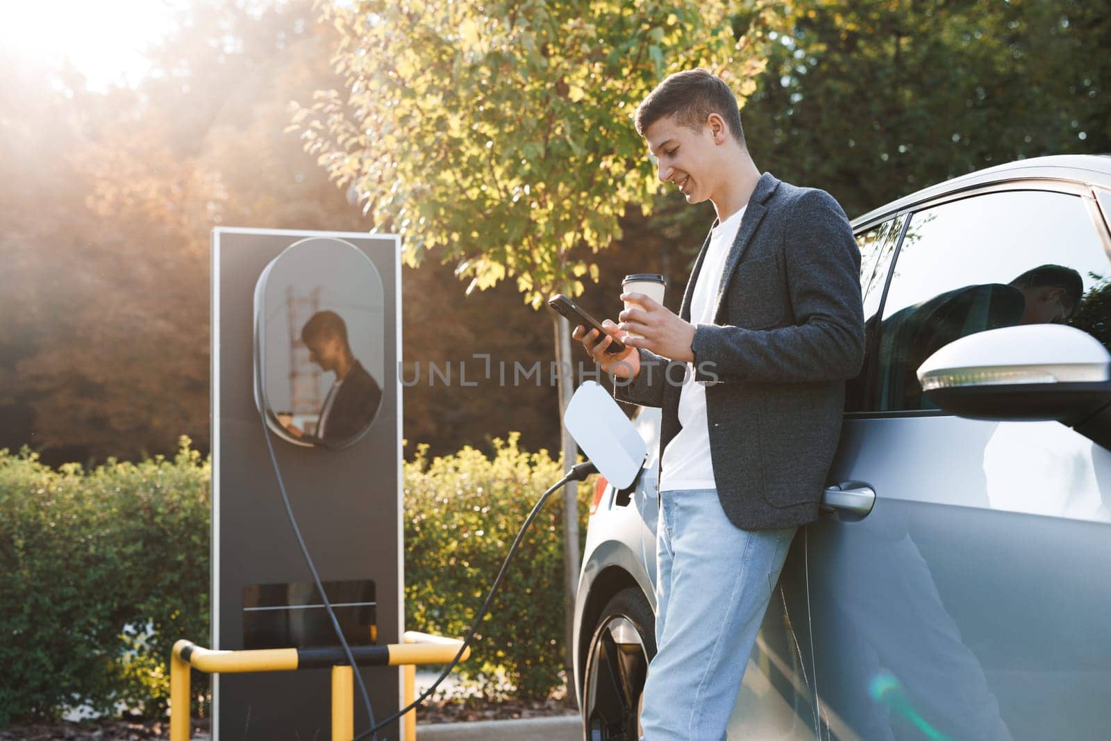 Caucasian businessman using smart phone and waiting power supply connect to electric vehicles for charging the battery in car. Plug charging an Electric car from charging station. Charging technology by uflypro