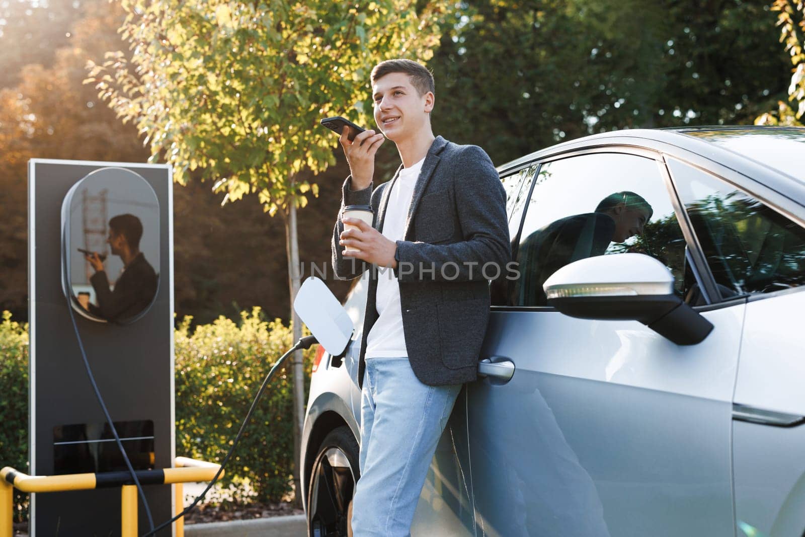 European man using smartphone voice messages and waiting power supply connect to electric vehicles for charging the battery in car. Plug charging an electric car from charging station by uflypro
