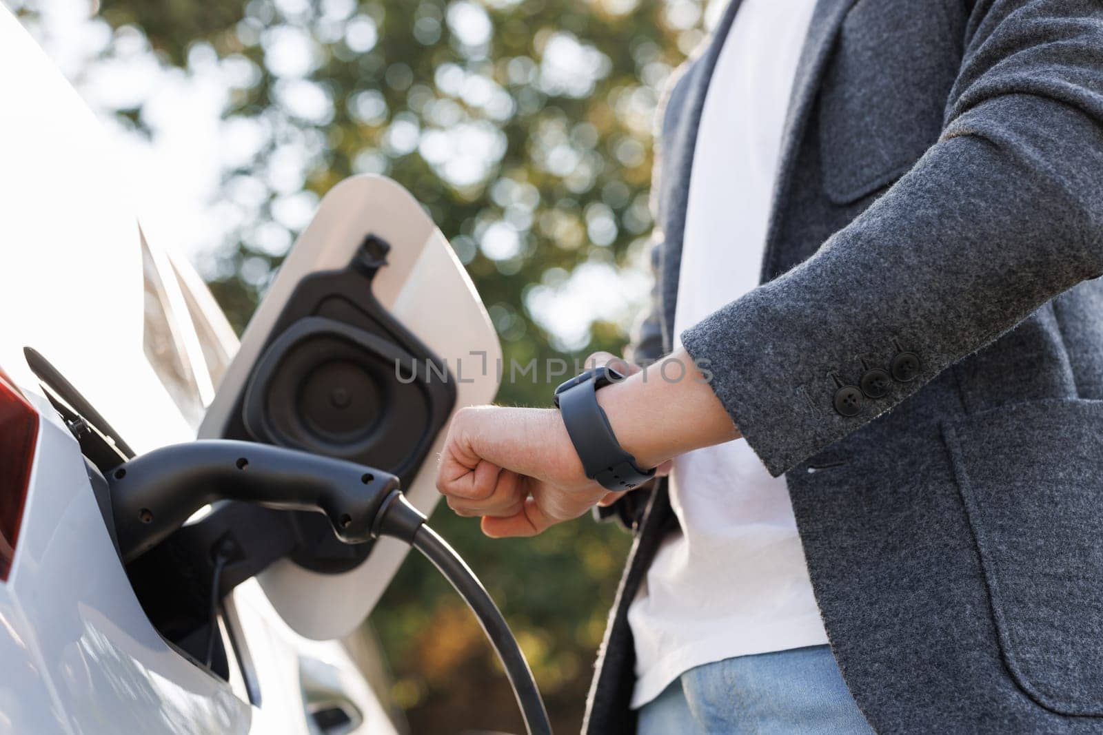 Male hand inserts power connector into EV car and charges batteries, uses smartwatch for activates start charging. Businessman plugging in charging cable to to electric vehicle.
