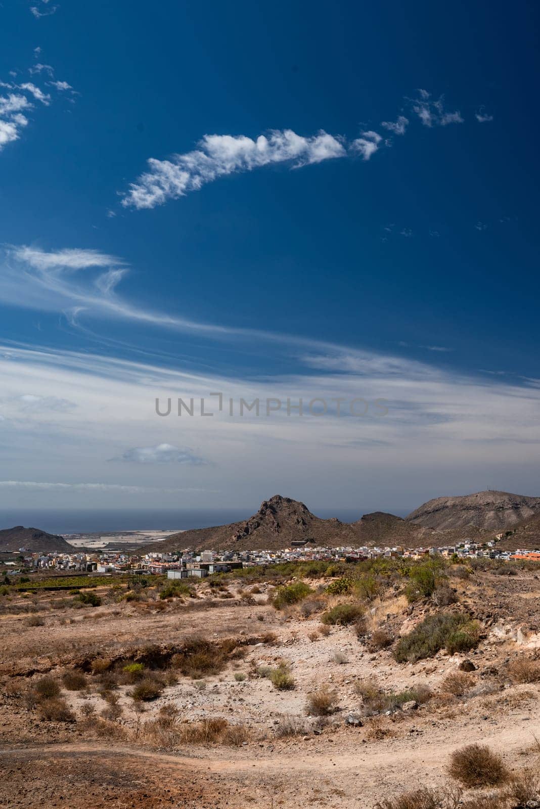 Hiking trail in a desert with a view of a seaside city, sea or ocean on a sunny day. Little mountains or hills at the horizon, blue sky and clouds. Little shrubs and a walking path between