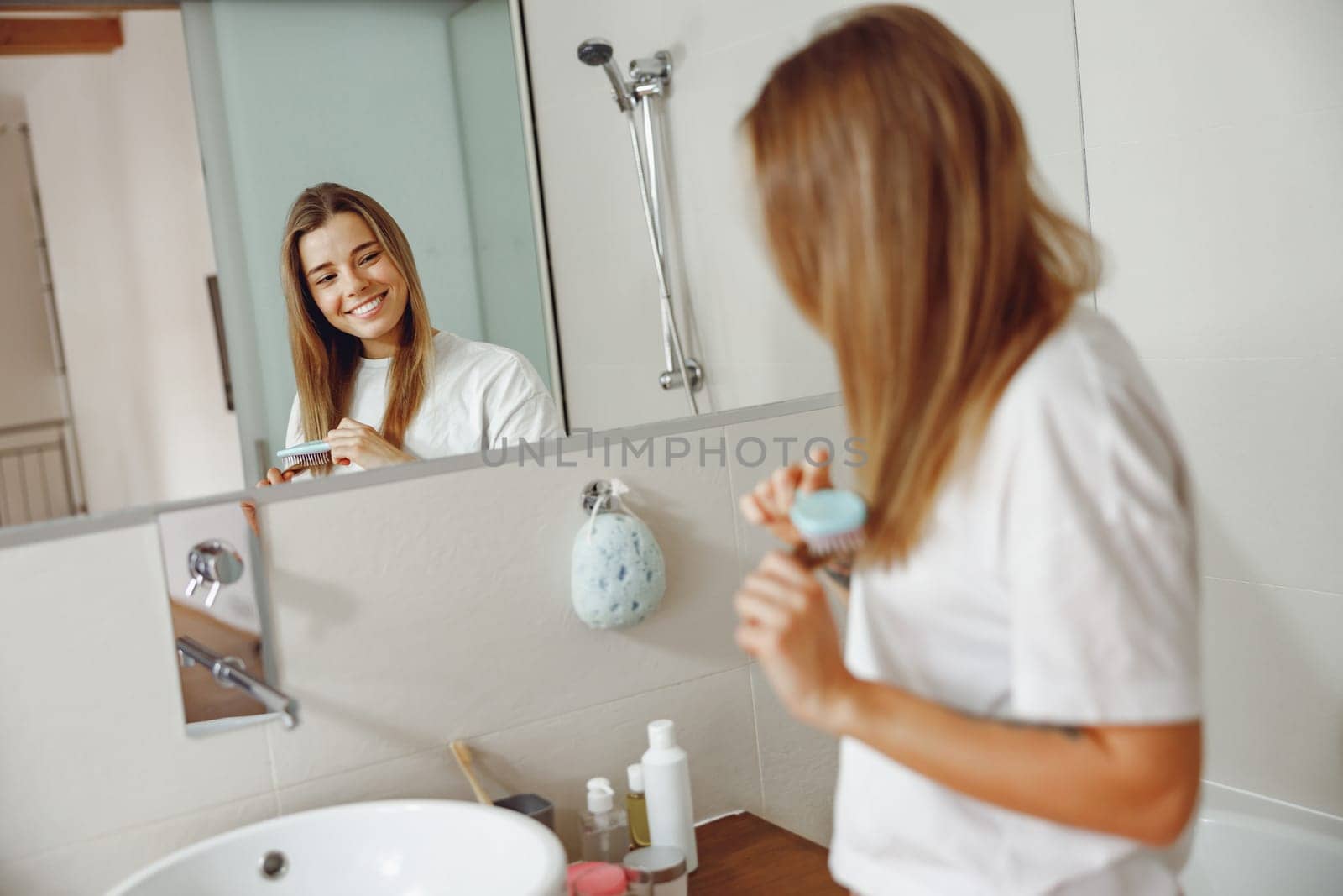 Woman standing in front of mirror at bathroom and combing her hair with brush after shower by Yaroslav_astakhov