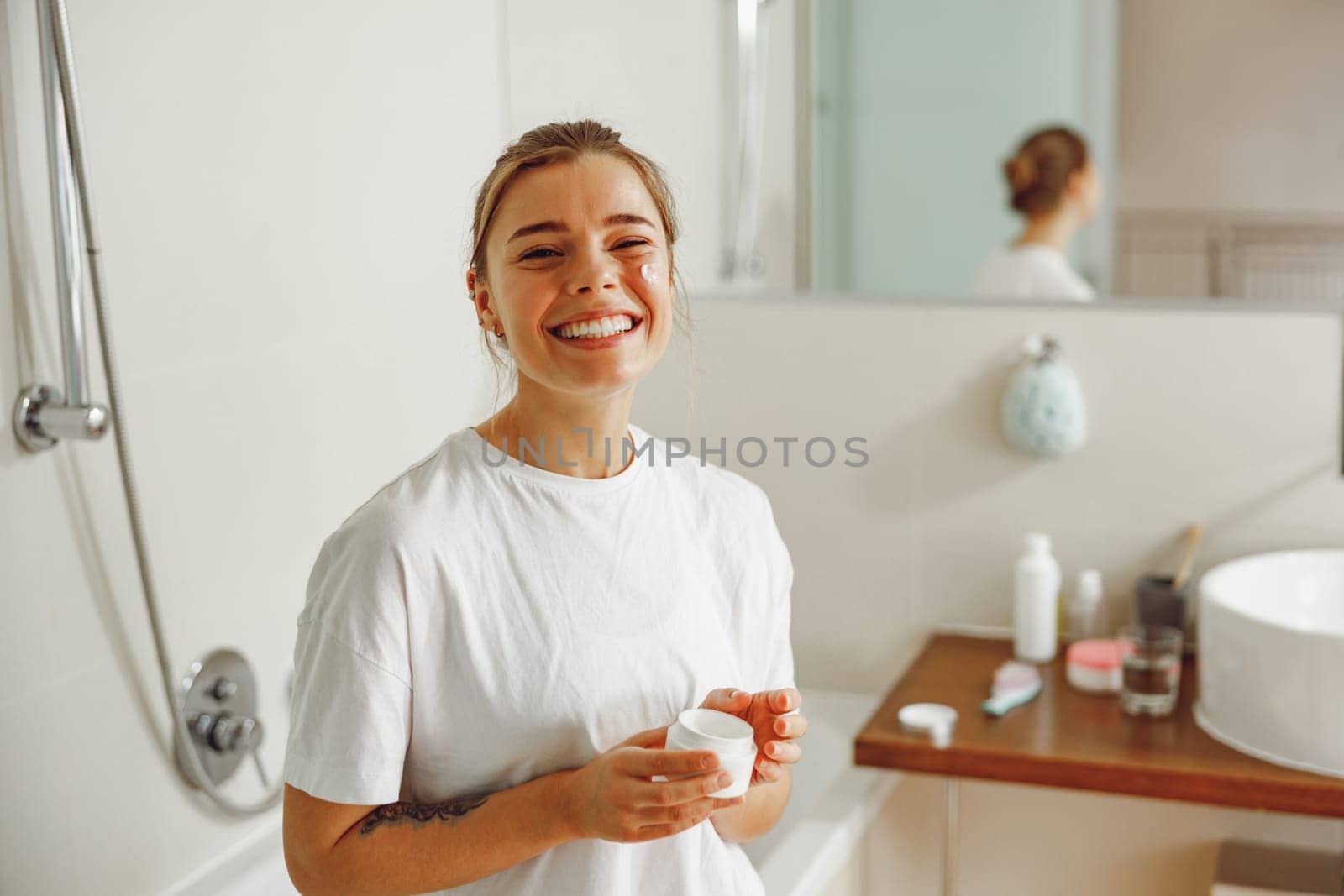 Beautiful young woman taking care of skin by applying moisturizer cream in bathroom