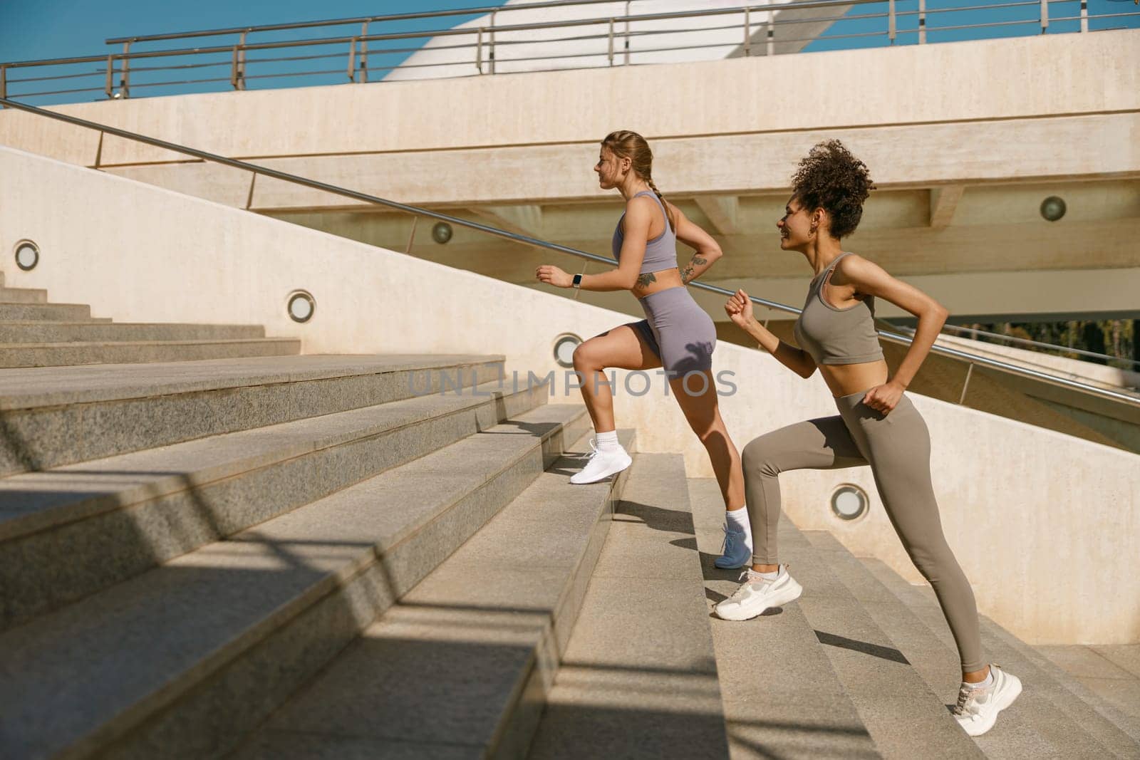 Two active female athlete friends in sportswear running on steps outdoors on a sunny day by Yaroslav_astakhov