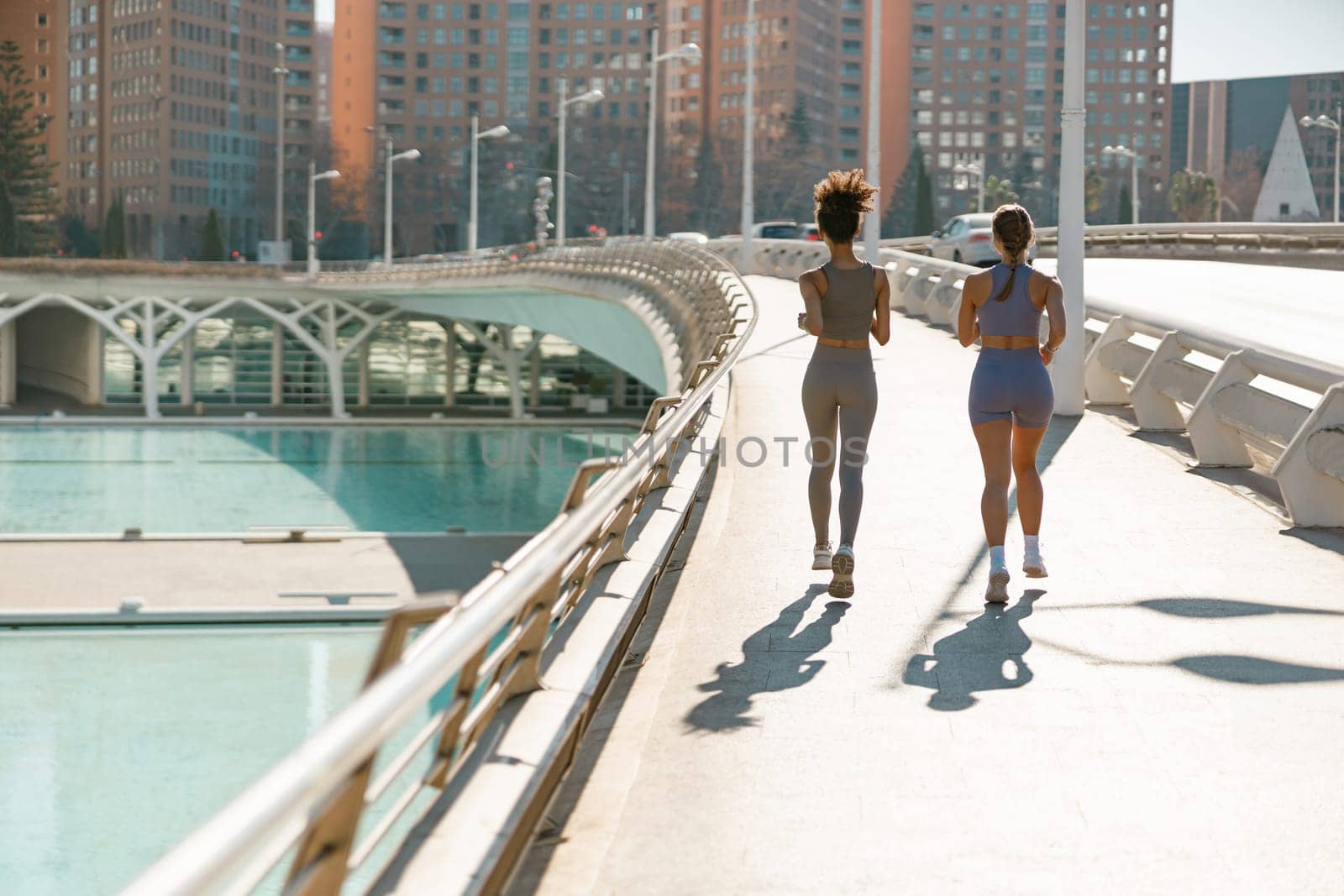 Back view of active women running side by side along the embankment on modern buildings background by Yaroslav_astakhov