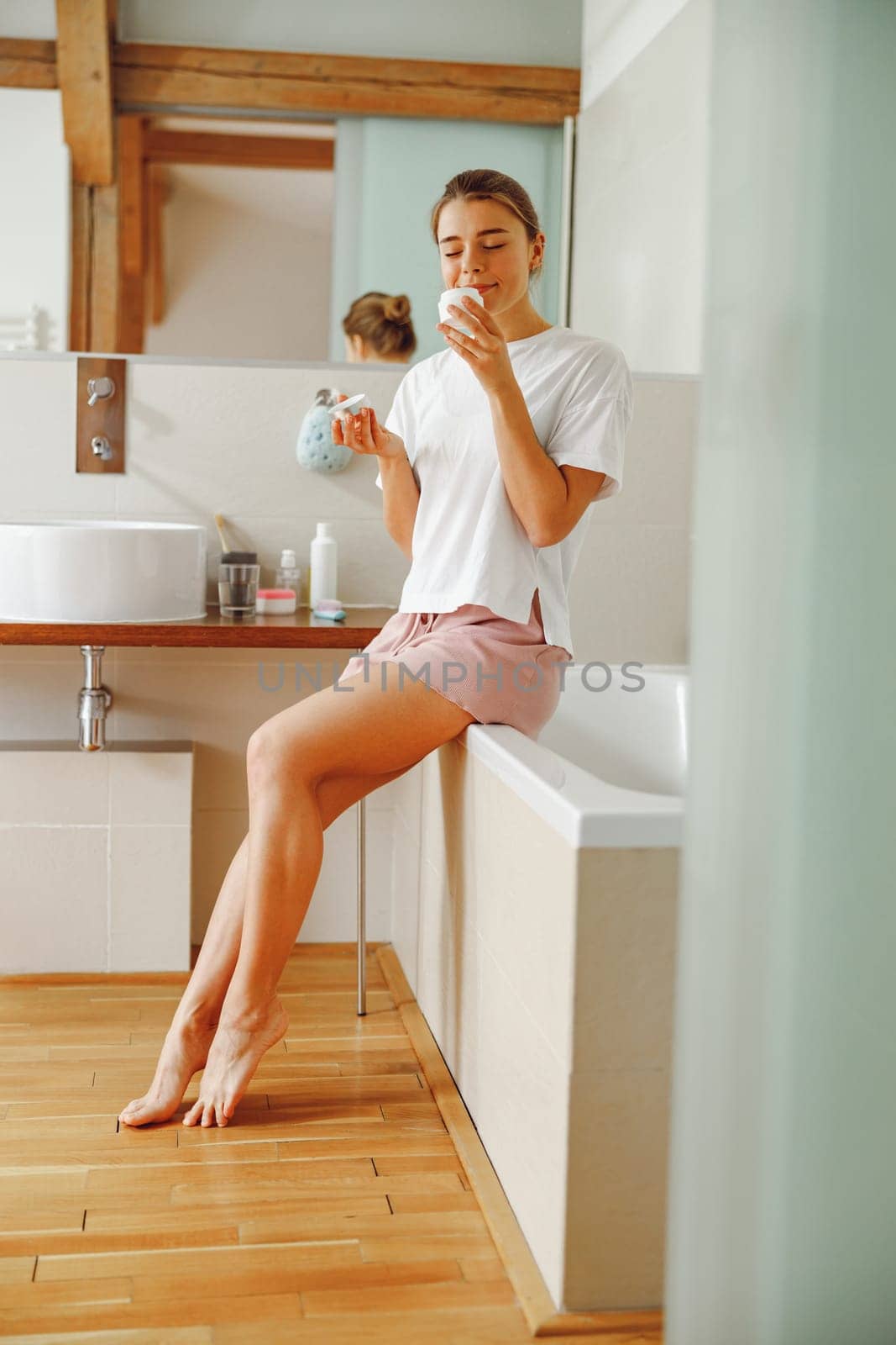 Young woman inhaling smell of jar with skincare cream sitting in bathroom. Home beauty routine by Yaroslav_astakhov