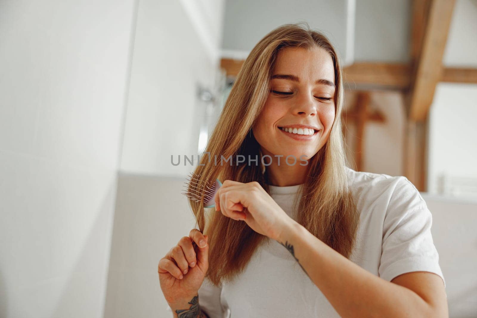 Woman standing in front of mirror at bathroom and combing her hair with brush after shower
