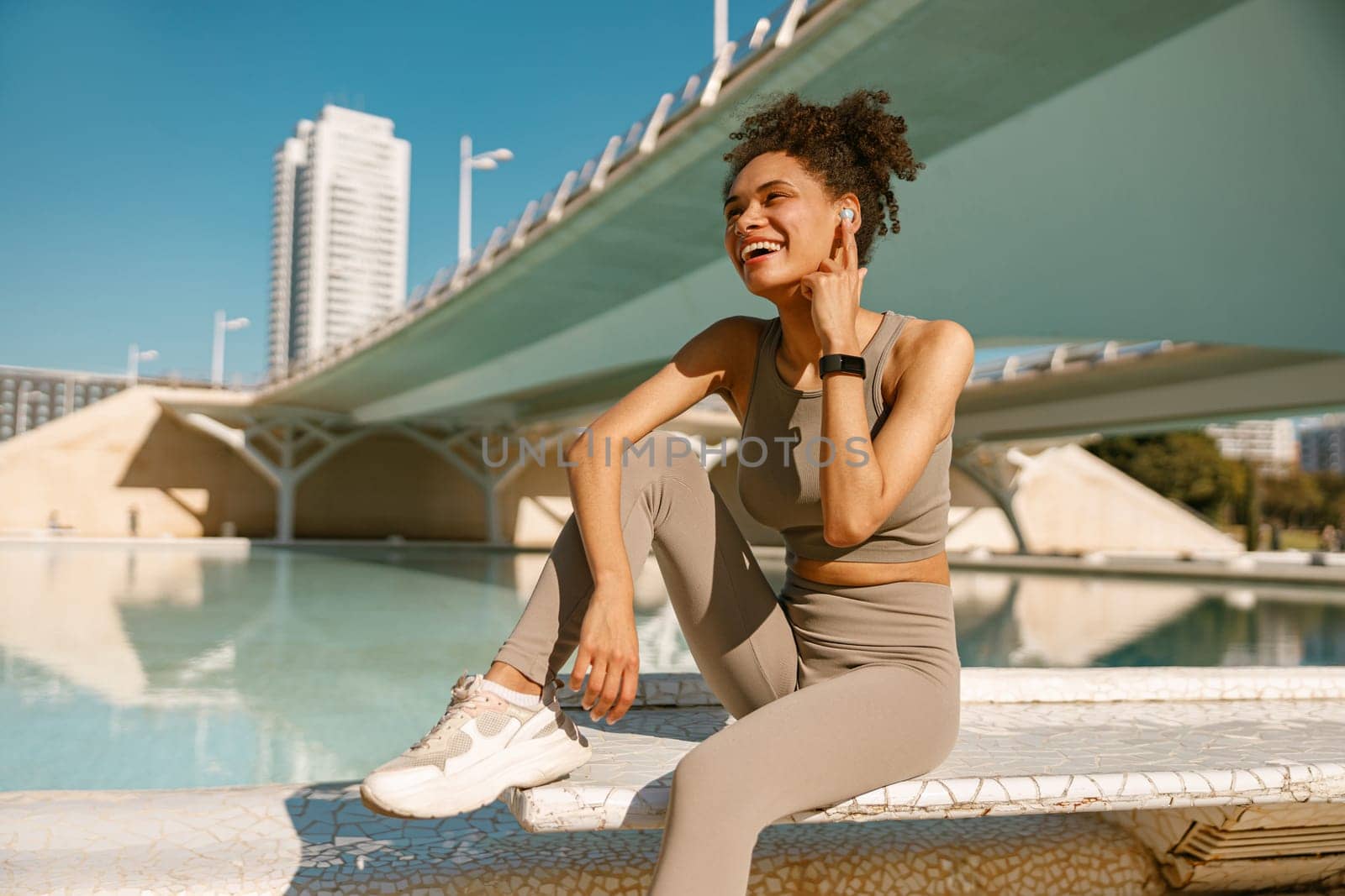 Smiling woman in sportswear have a rest after workout outside sitting on bridge background by Yaroslav_astakhov