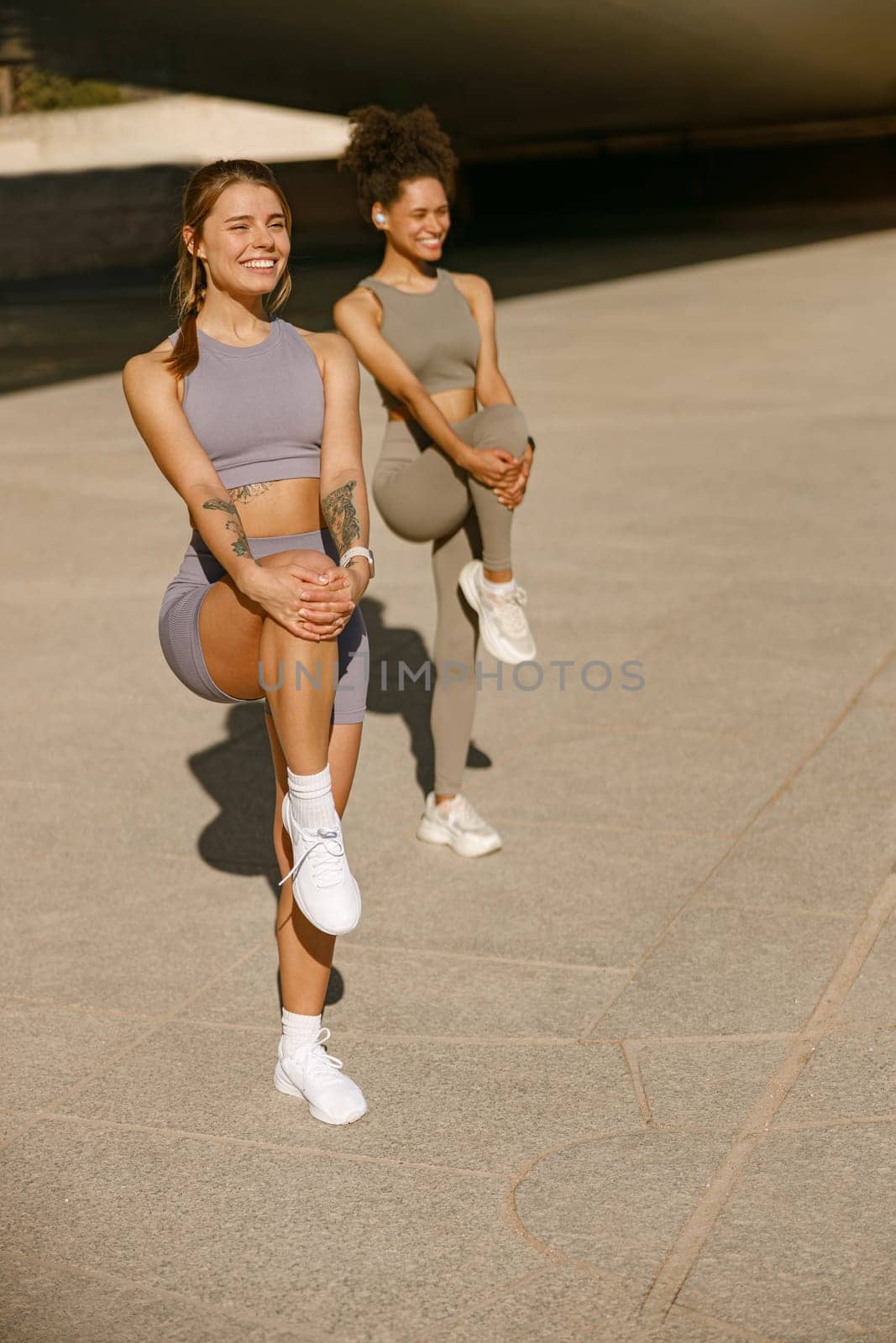 Two young sporty women in tracksuits are stretches legs outside on modern building background by Yaroslav_astakhov