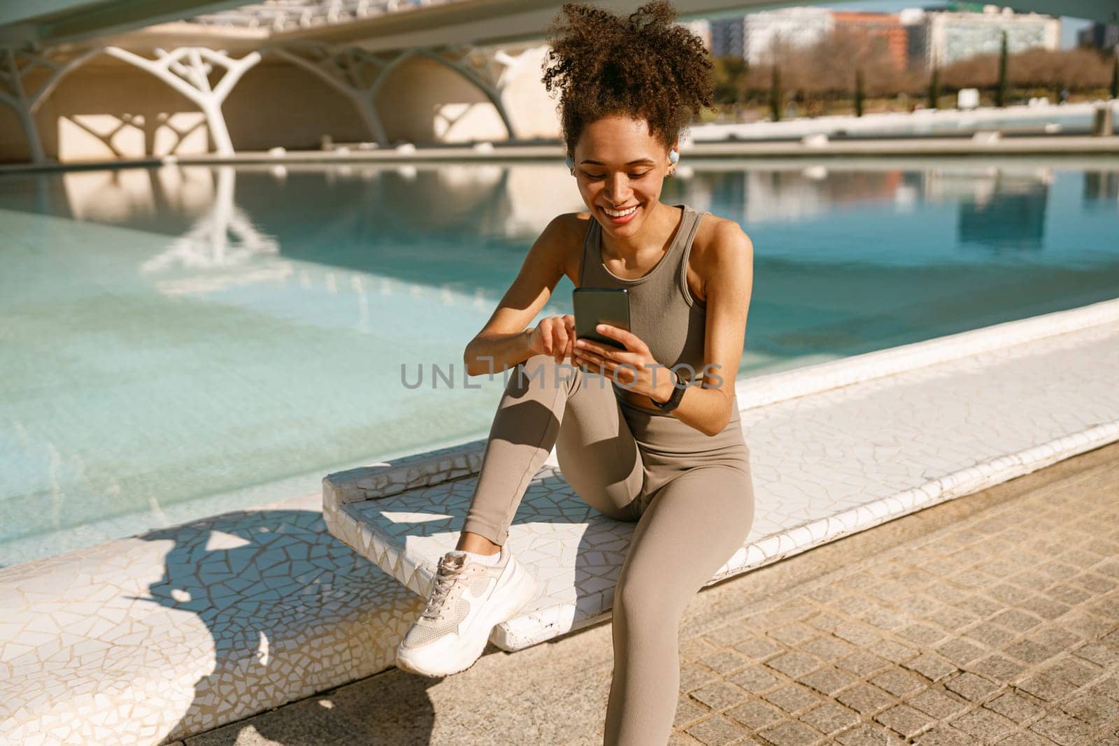 Smiling woman in sportswear sitting outdoors after training and use mobile phone by Yaroslav_astakhov