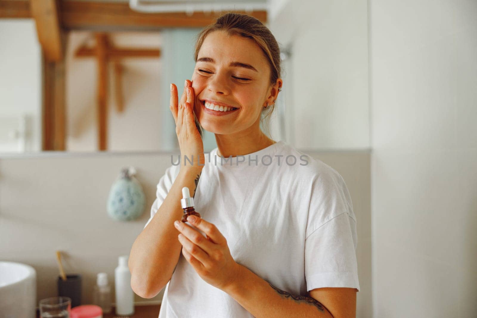 Smiling young woman applying cosmetic serum onto her face in bathroom. Facial Skincare by Yaroslav_astakhov