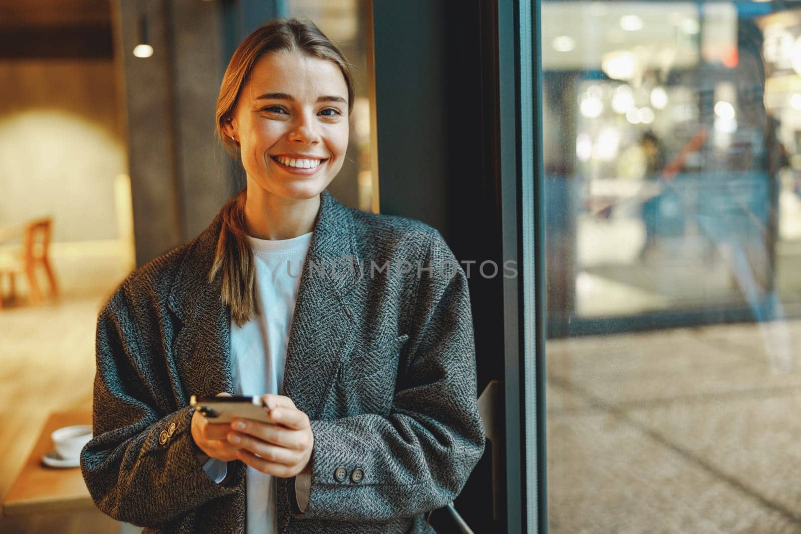 Young female entrepreneur is use phone while standing in cafe near window. Distance work concept by Yaroslav_astakhov