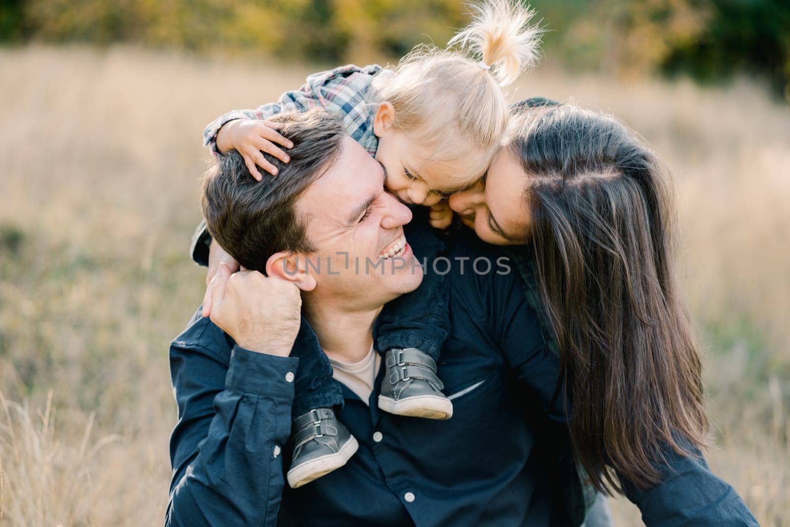Mom with a little girl on the shoulders of dad kiss him, hugging from behind. High quality photo