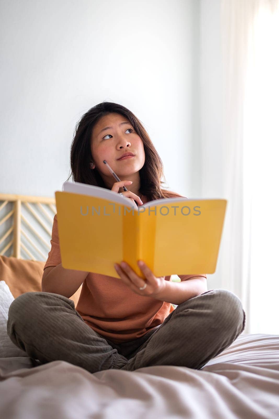 Teen asian girl sitting on bed thinking, using diary and pen to write down thoughts and ideas. Vertical. by Hoverstock