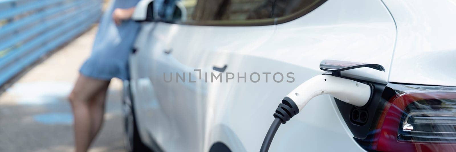 Electric vehicle recharging battery from home EV charging station using alternative energy with net zero emission on blurred background of young girl charging before vacation travel.Panorama Perpetual