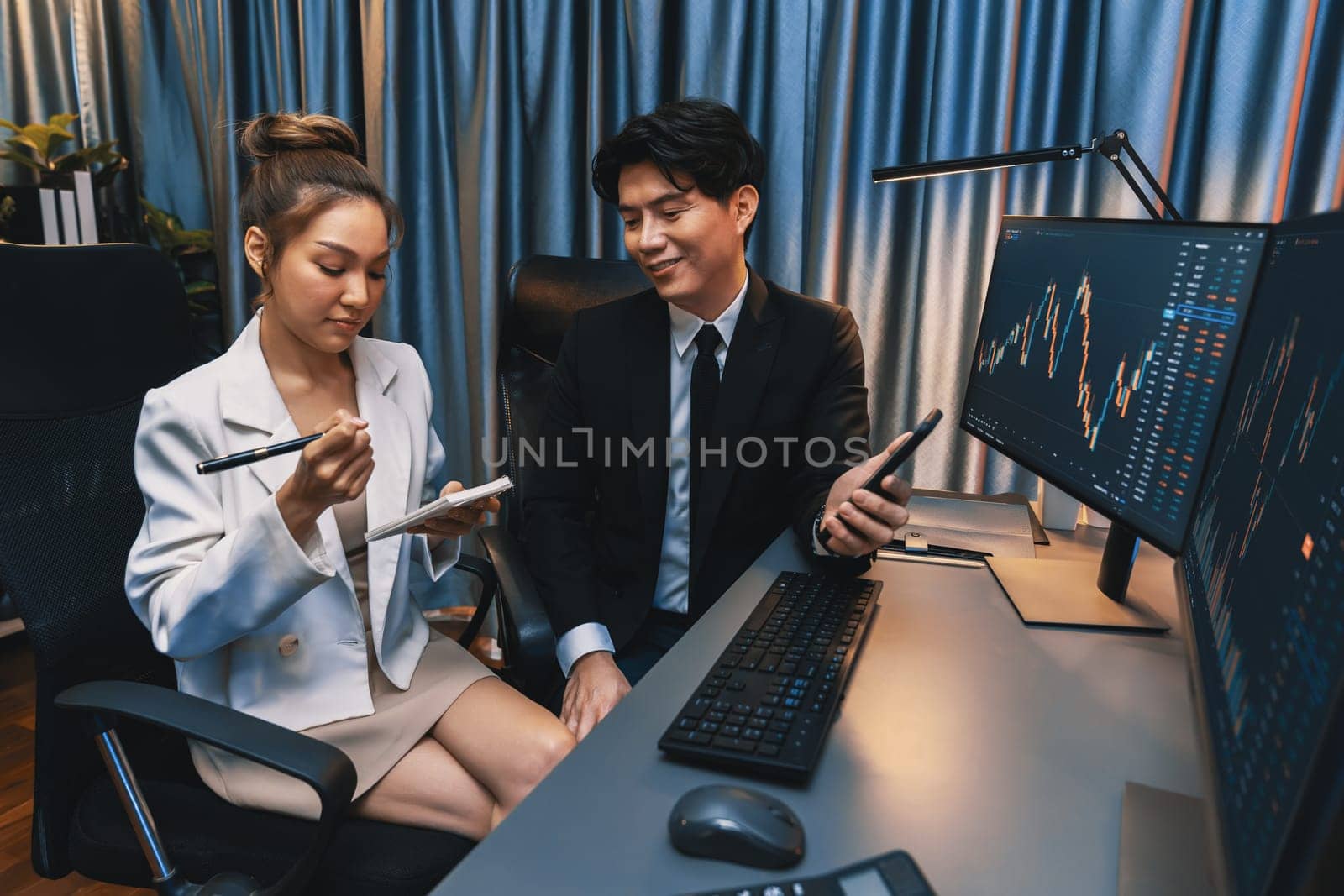 Asian businessman showing stock market application on mobile phone to partner pointing by pen, comparing financial digital investment database on computer screen at neon light at night time. Infobahn.