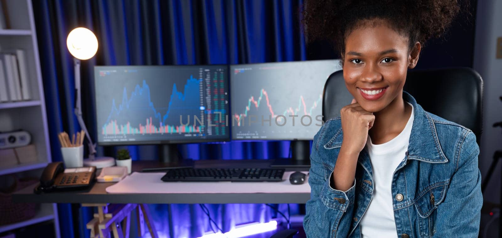 Profile of young African American businesswoman smiling on happy face wearing jeans shirt, sitting on chair against stock exchange market screen background. Concept of investment blogger. Tastemaker.