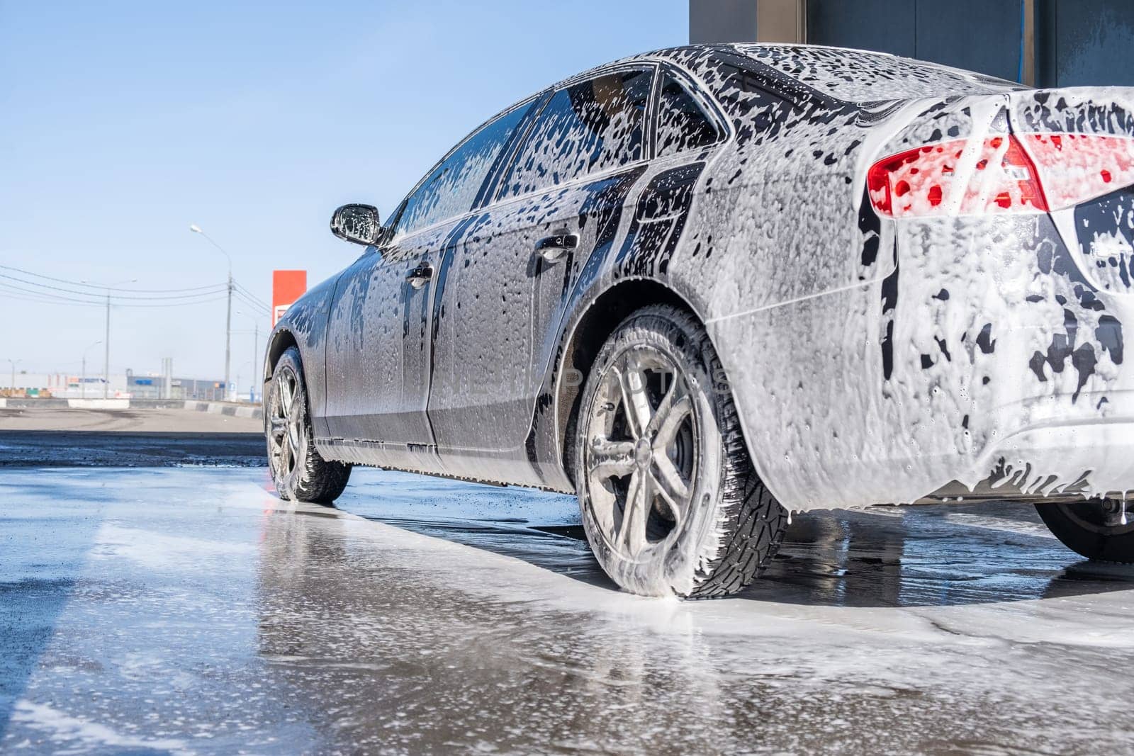 A vehicle is engulfed in soapy foam during a car wash, covering the tires, wheels, hood, and entire exterior of the car. Self-service car wash