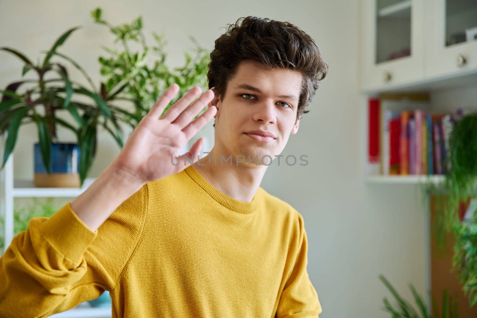 Portrait of young handsome guy waving hand looking at camera, in home interior. Smiling confident male 19-20 years old in casual yellow with curly hairstyle looking at camera. Lifestyle, youth concept