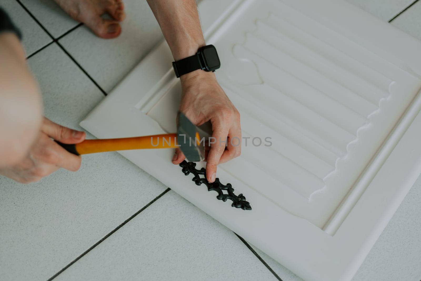 Hands of a young Caucasian unrecognizable man hammers a nail, installing an antique carved black lock on a white cabinet door, squatting on a tiled floor, close-up top view.