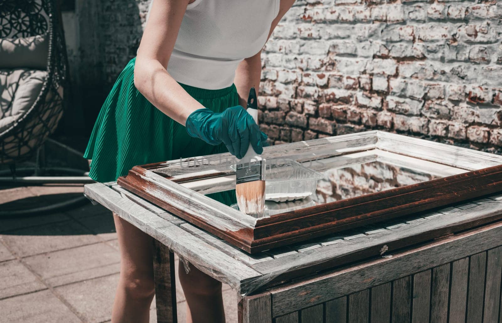 One young Caucasian unrecognizable girl in green shorts and gloves paints a mirror frame with white paint with a brush, standing in the backyard of the house on a summer day, close-up side view.