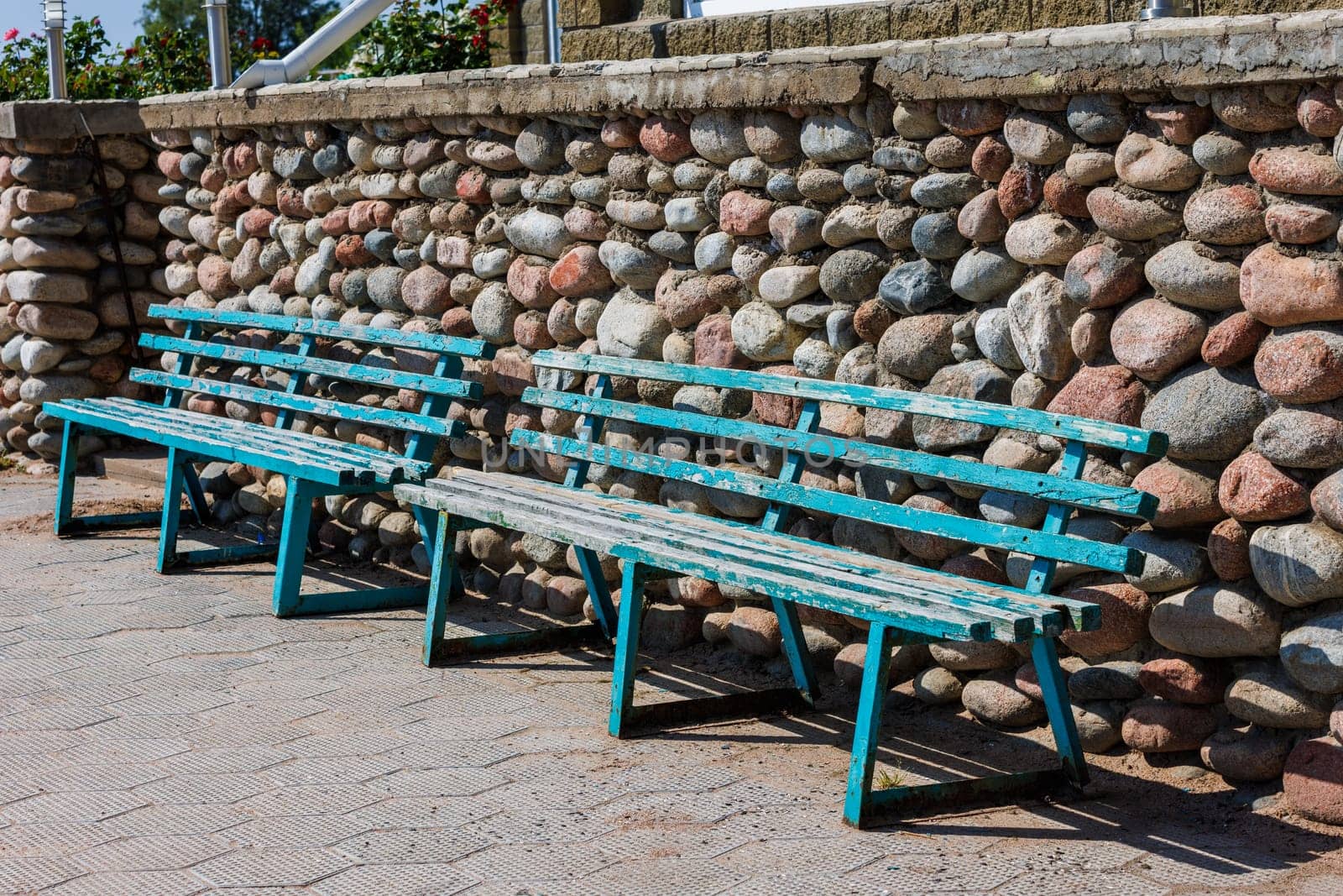 Two vacant outdoor wooden benches with peeled blue paint lined up against a stone wall at sunny summer day.