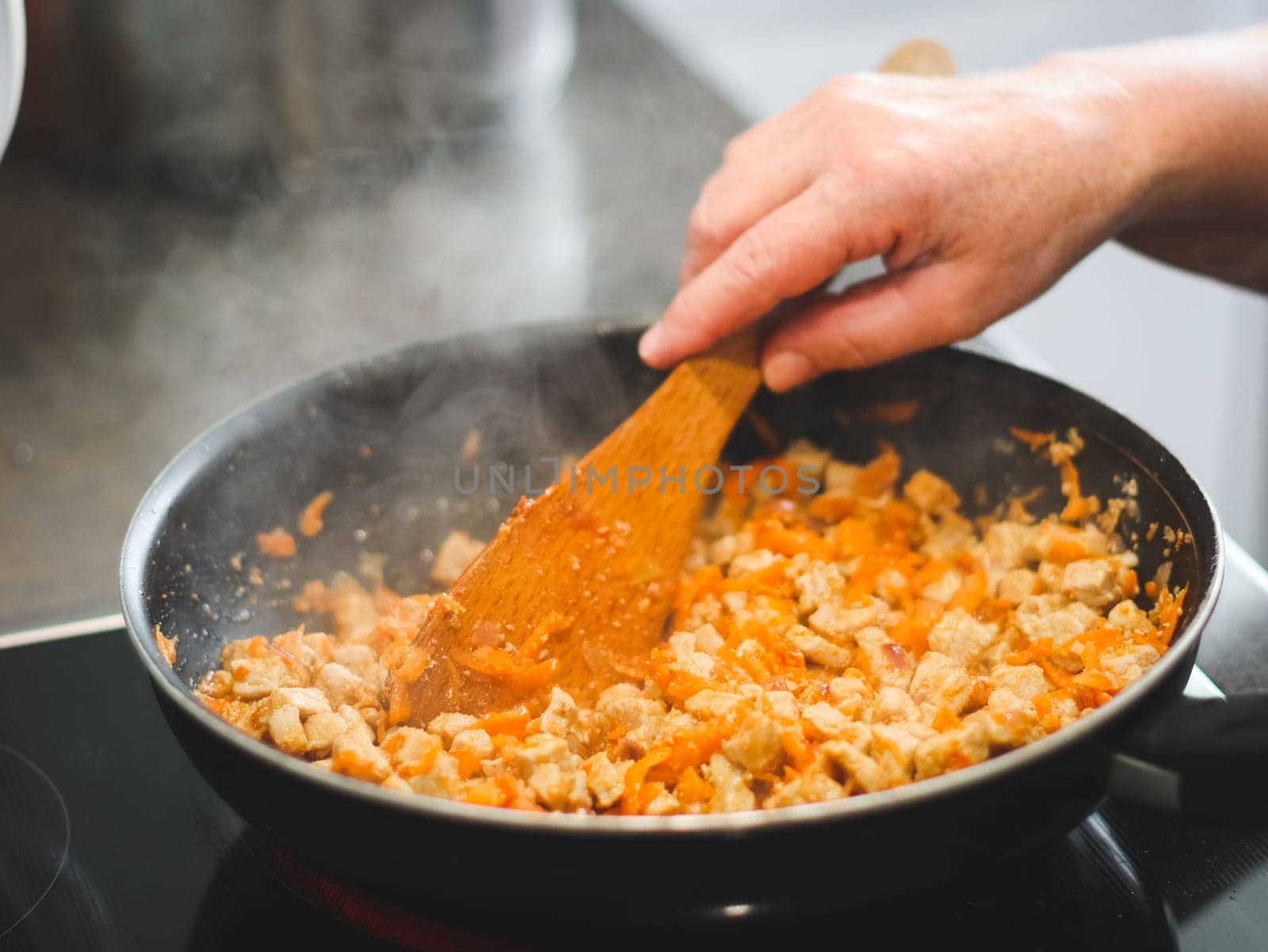 The hand of a senior woman holds a wooden spoon and stirs meat with carrots that are fried in a frying pan on an electric stove, close-up side view. Concept step by step instruction, cooking home, traditional recipes, national cuisine, cabbage rolls, dolma.