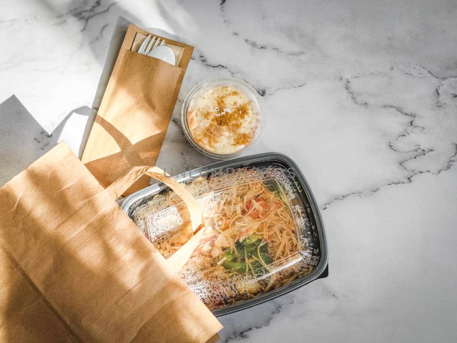 Ready-made noodles with vegetables in a black container, tiramisu dessert and a spoon with a fork in cardboard spilling out of a paper bag lie on a marble table, flat lay close-up. Asian food concept.