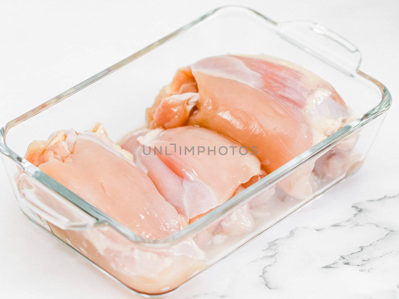 Raw boneless chicken thigh meat lies in a glass baking dish on a white marble table, close-up side view. Concept of raw chicken meat, raw food.