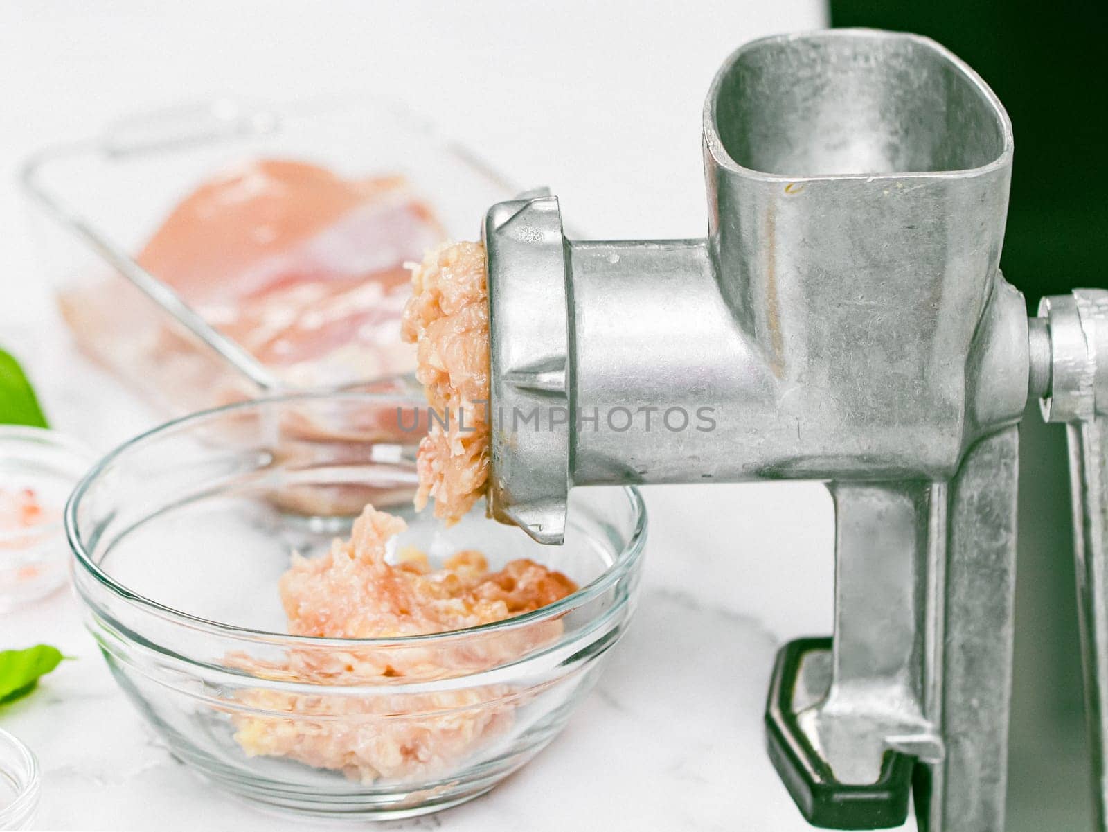 Manual retro meat grinder with minced meat in a glass bowl with boneless chicken thighs and basil leaves on a marble table, close-up side view. The concept of home cooking and healthy eating.