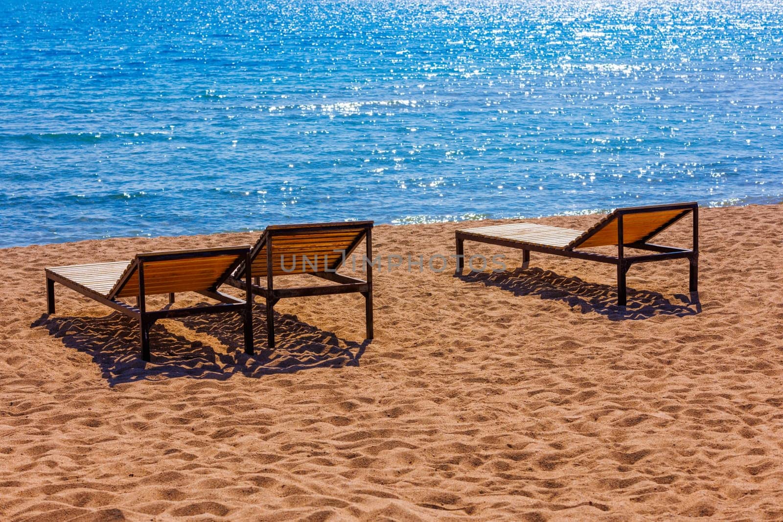 Three empty outdoor lounge chairs on sandy beach near blue water at sunny off-season day.