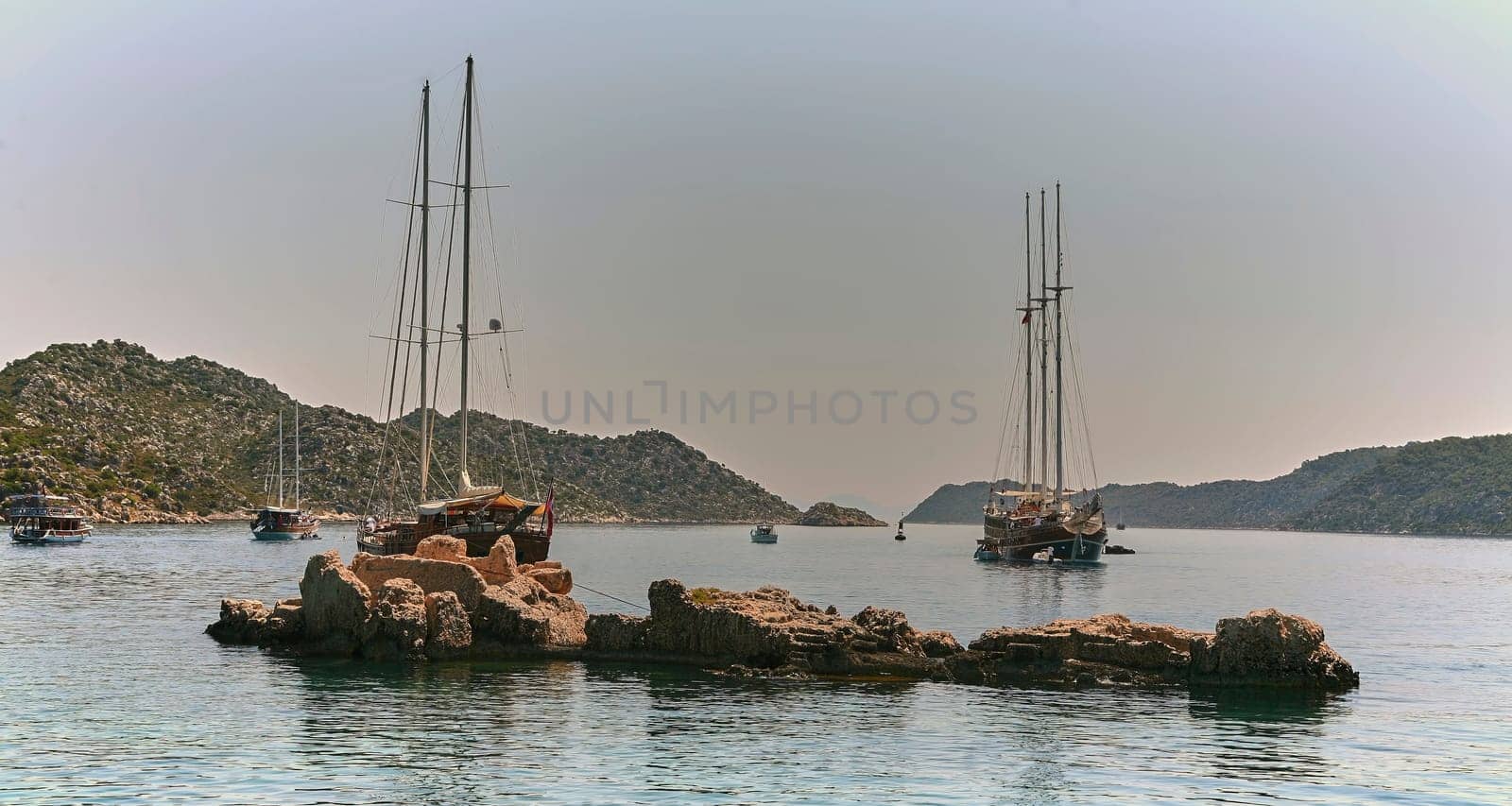 Wooden sailing boat in calm sea with green mountains. Two masts, white sails. Clear blue-green water, blue sky, white clouds. Marina with more boats. Great for travel brochure.