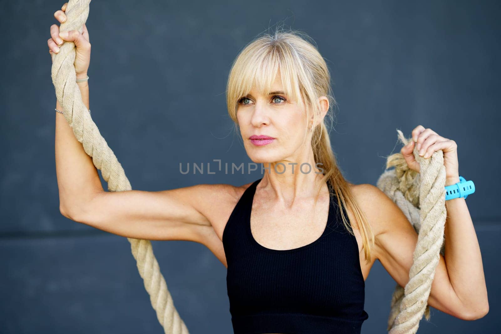 Young fit female wearing sportswear holding rope against gray background in fitness studio looking away thoughtfully while exercising at modern gym