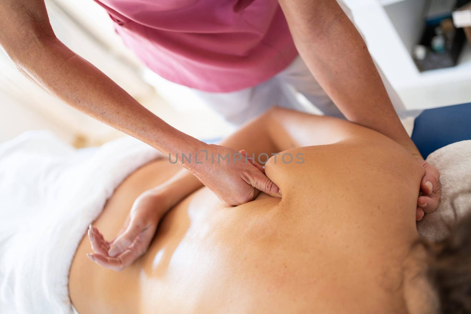 High angle of anonymous osteopathic therapist massaging using fist on shoulder bone at back of patient lying face down during physiotherapy session in modern clinic