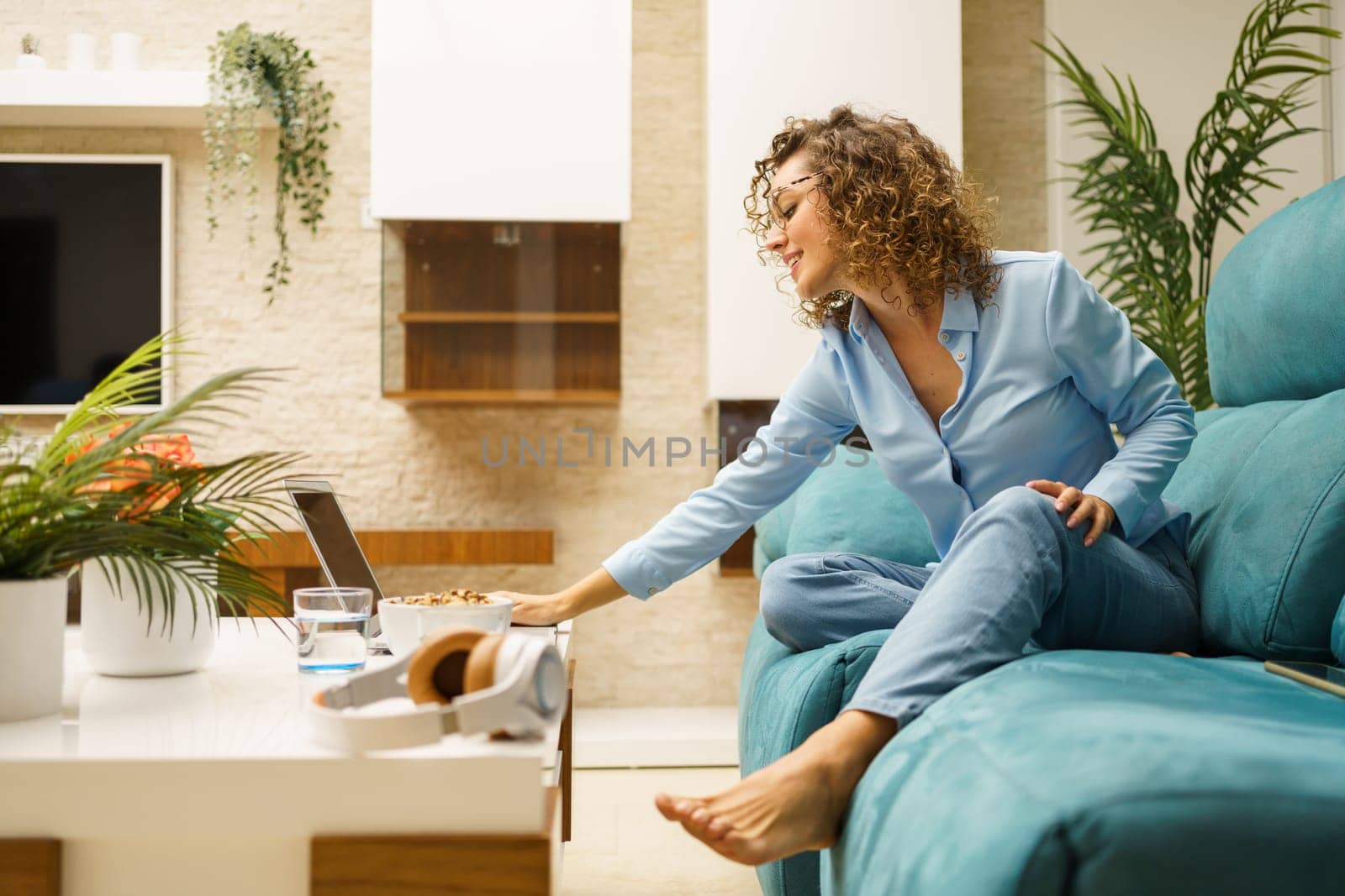 Young positive woman sitting on sofa and using laptop during breakfast at home by javiindy