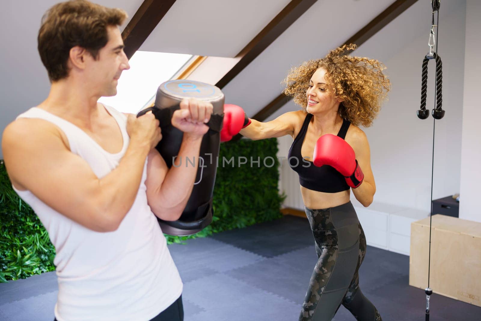 Cheerful young curly haired female athlete in sportswear and red boxing gloves punching bag during training with personal instructors assistance