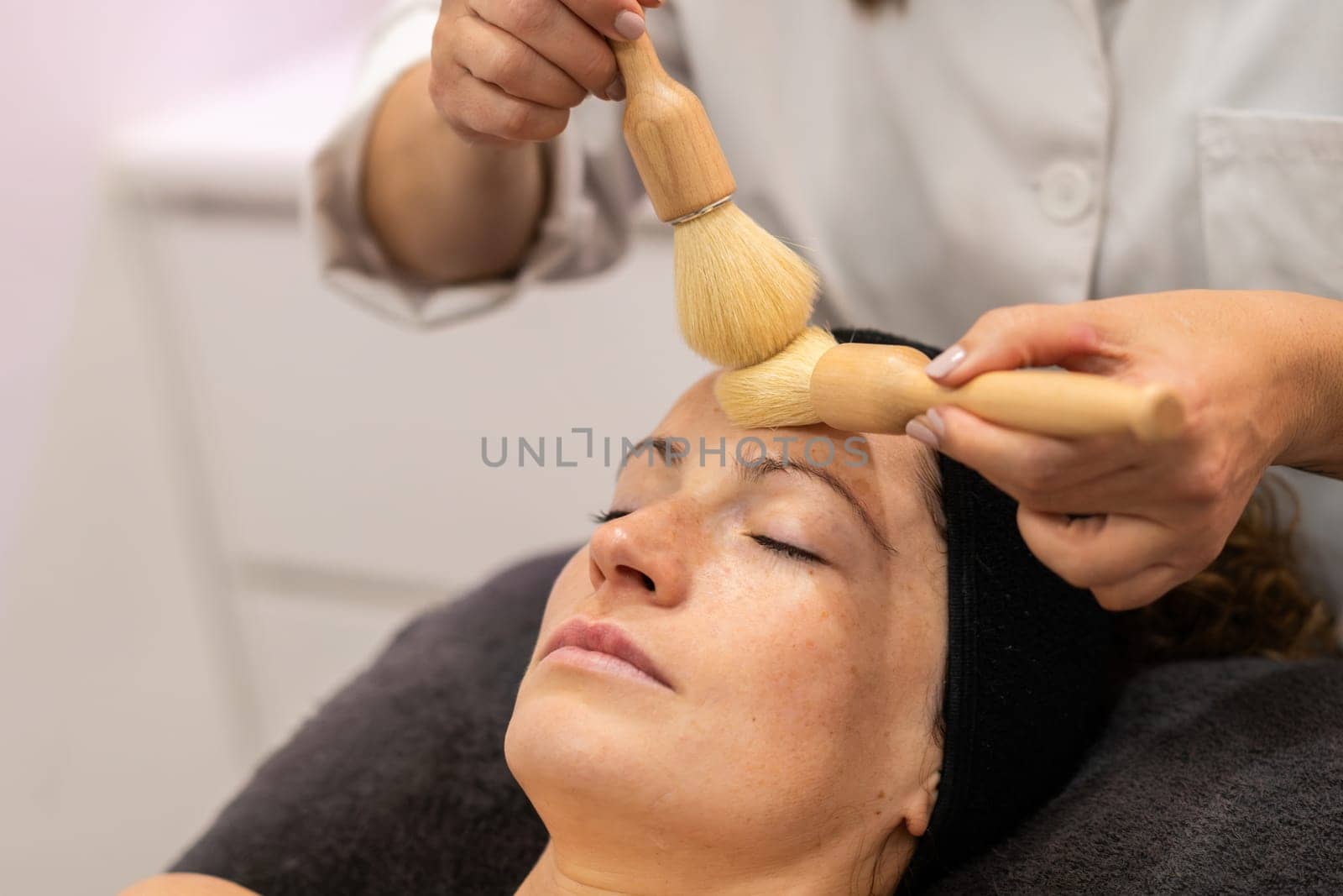 Crop beautician applying cosmetic product on face of female with brushes in beauty salon by javiindy