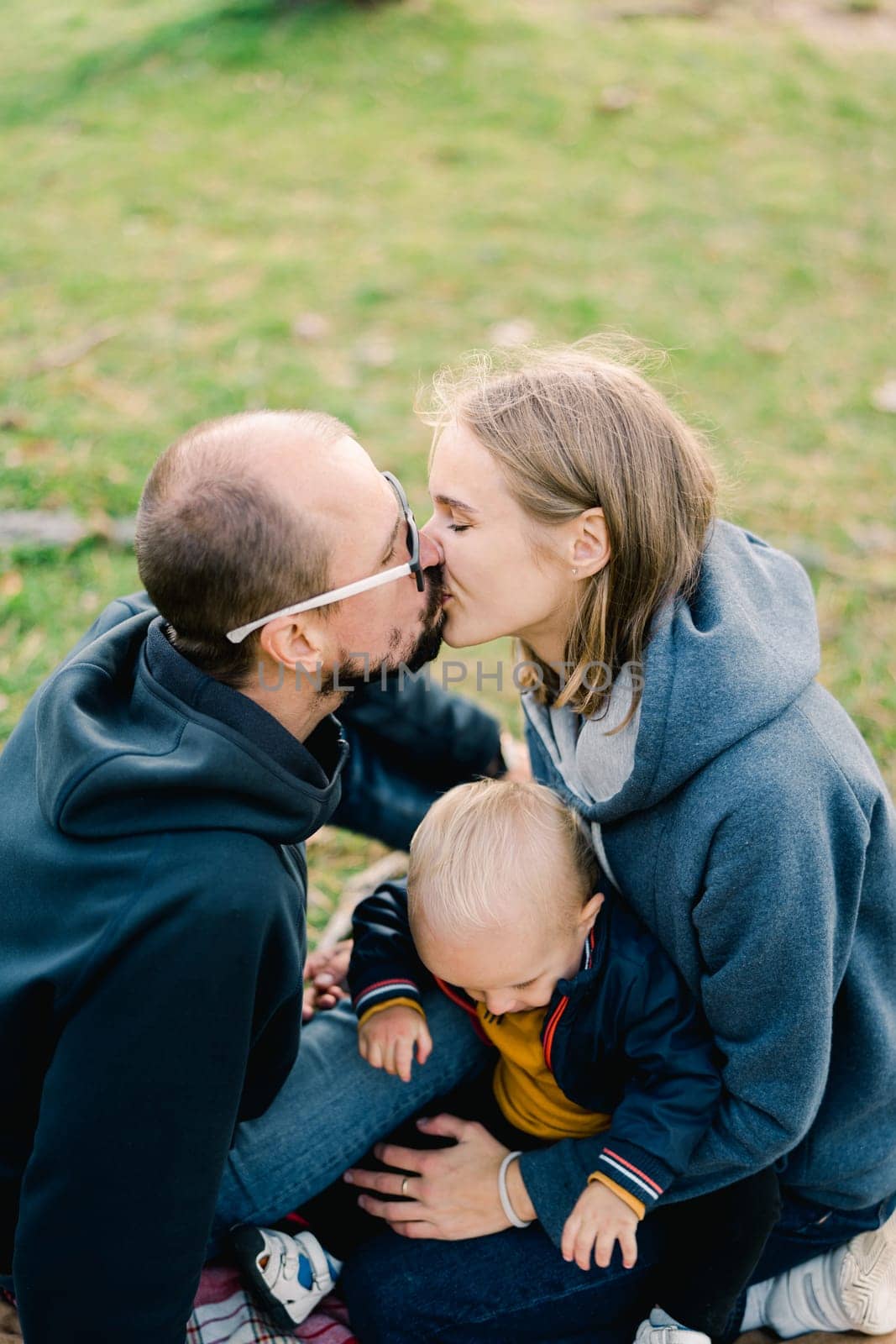Dad kisses mom with a little boy on her lap while sitting on a green lawn. High quality photo