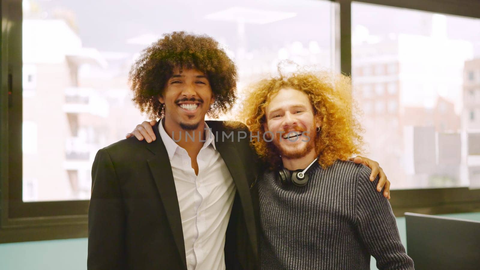 Two coworkers with curly hair embracing and smiling at camera in the office
