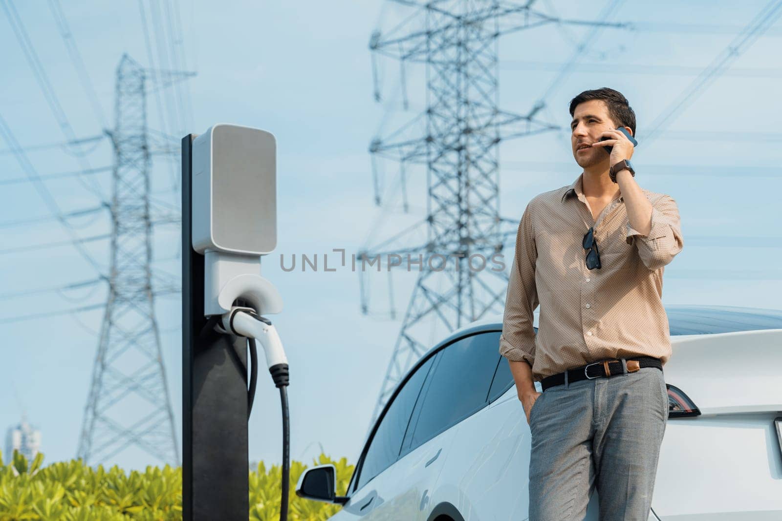 Man talking on the phone while recharge EV car battery at charging station connected to power grid tower electrical as electrical industry for eco friendly car utilization.Expedient