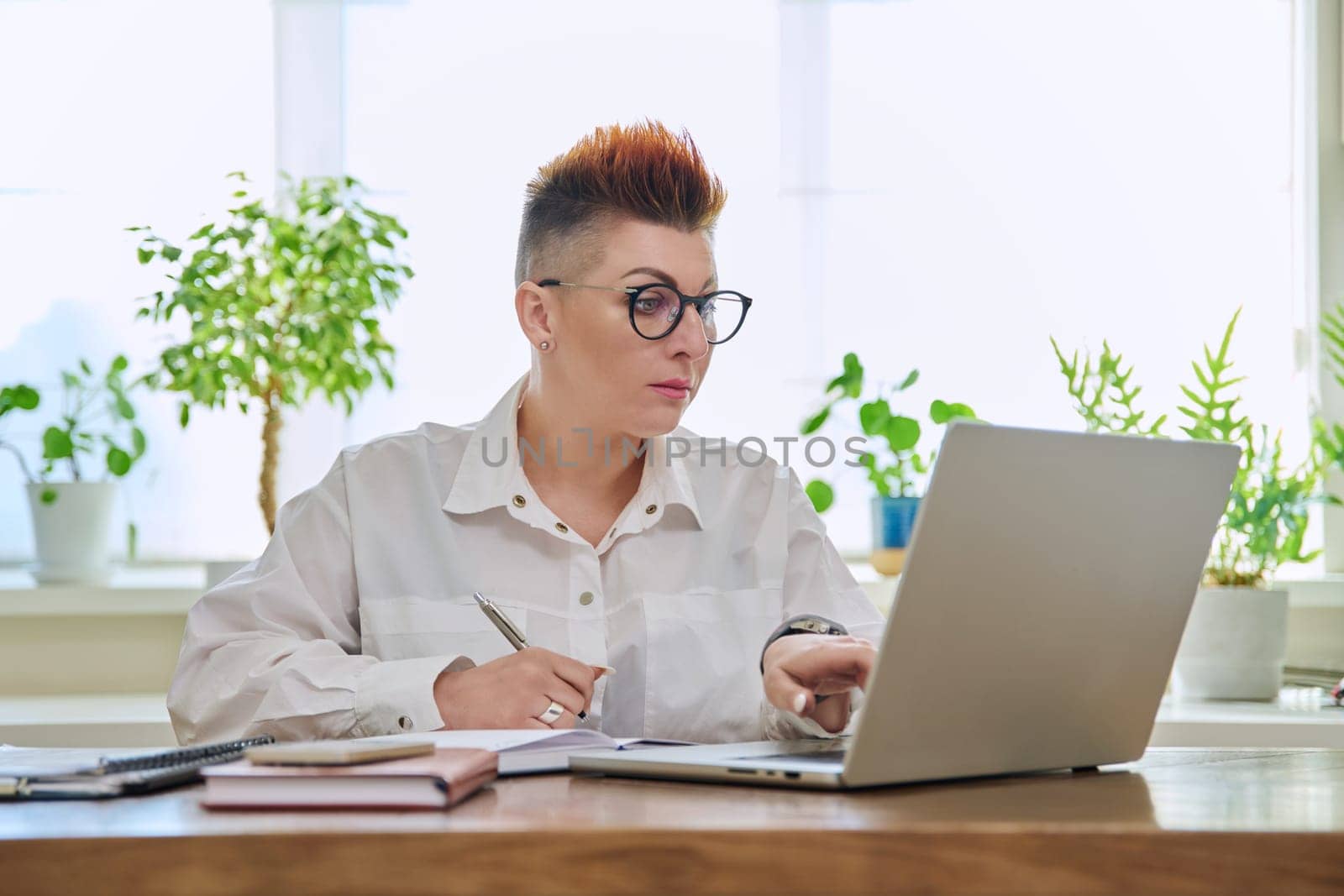 Middle-aged serious woman working at computer laptop in home office. Mature female sitting at desk typing and looking at monitor. Remote business, teaching, work, blogging, freelance