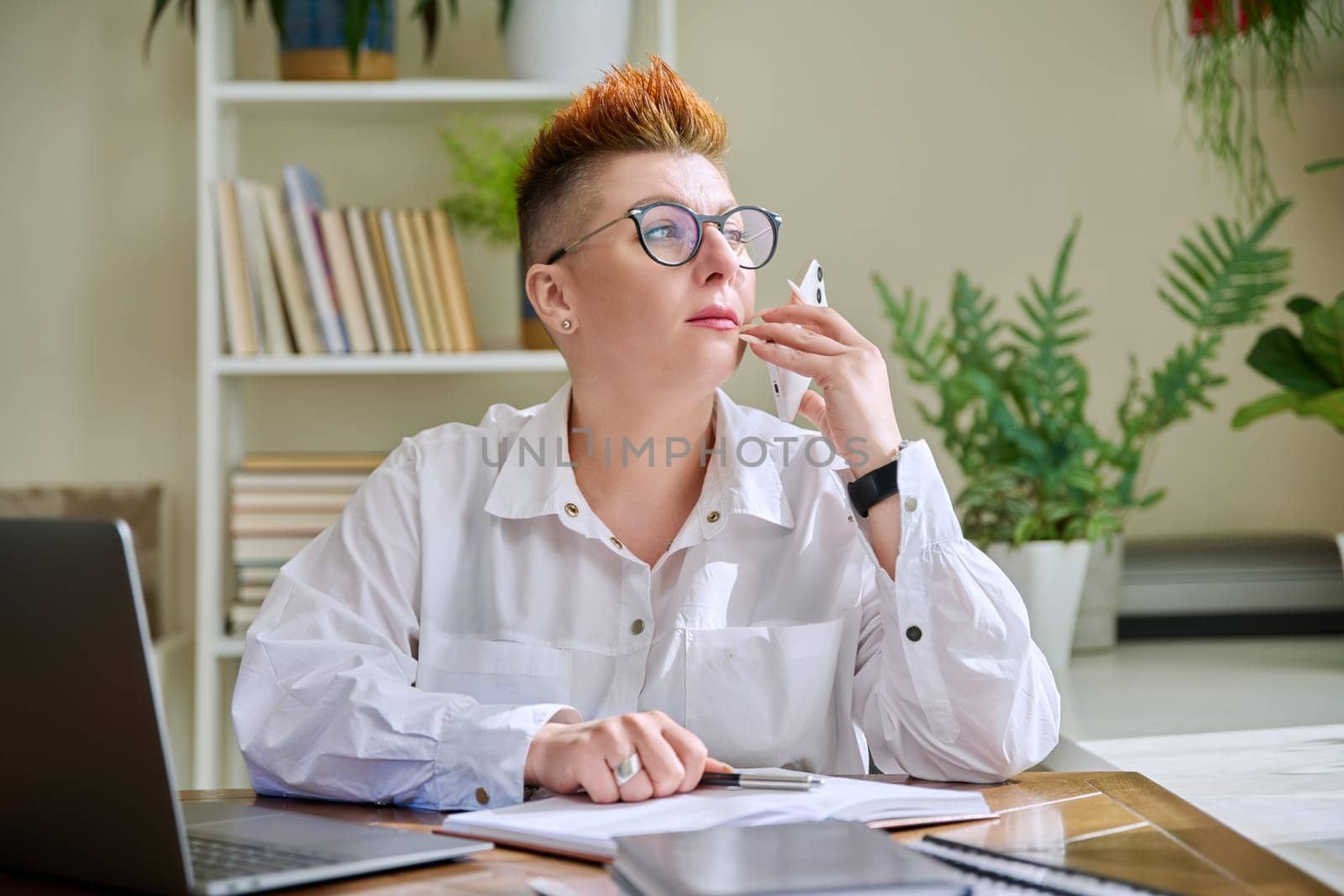 Middle-aged business woman talking on mobile phone sitting at desk with laptop computer. Serious confident mature female working with business papers. Work, workplace, 40s people