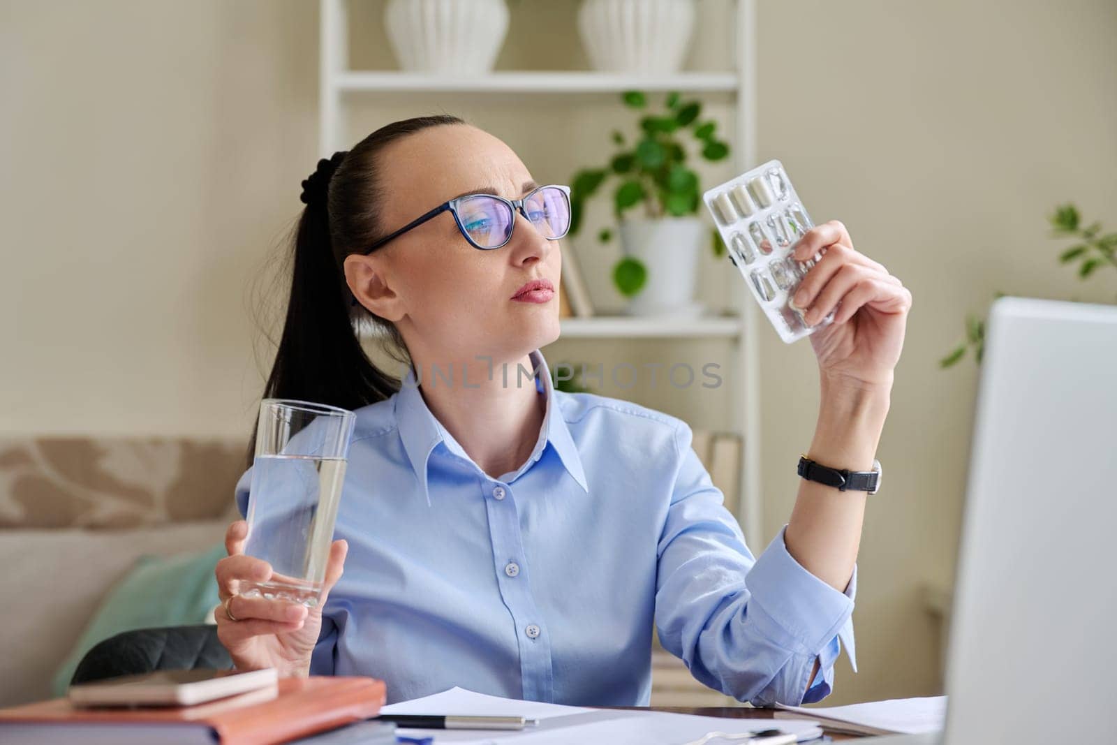 Serious woman at workplace holding blister with capsules glass of water by VH-studio